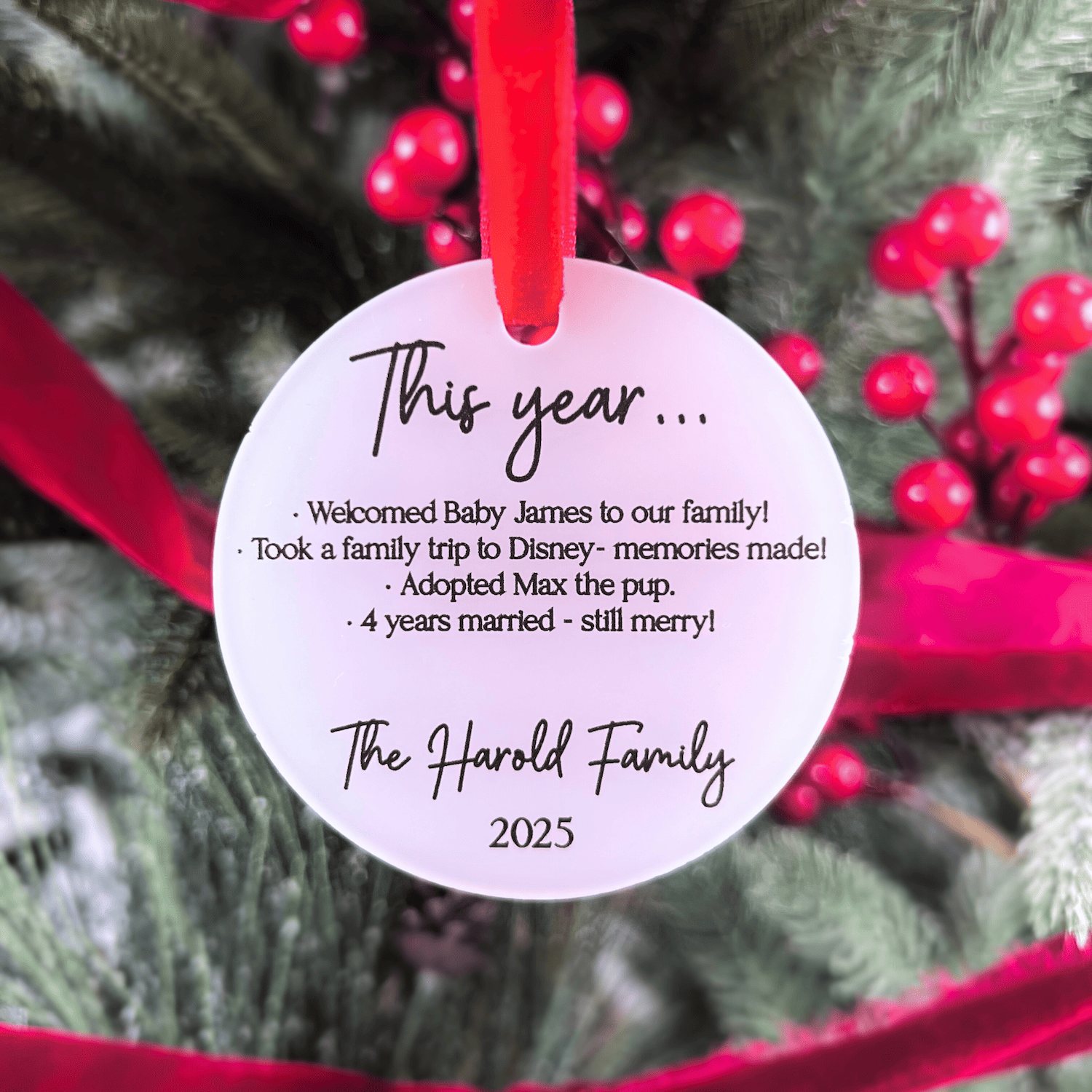White acrylic ornament with red ribbon hanging on festive tree, personalized with "This year..." heading, family milestones listed, and "The Harold Family 2025" at bottom.