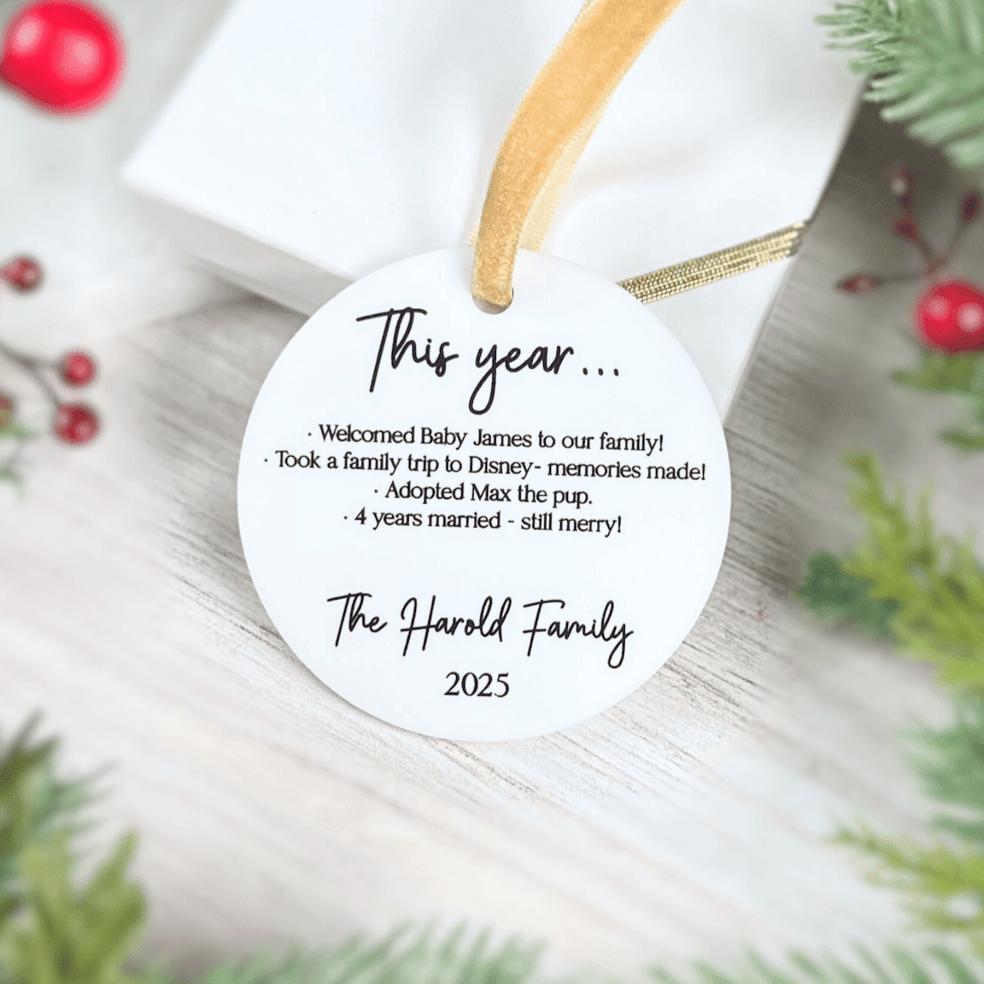 White round acrylic ornament with gold string showing "This year..." at top, family highlights text, "The Harold Family 2025" at bottom, surrounded by evergreen branches and red berries.
