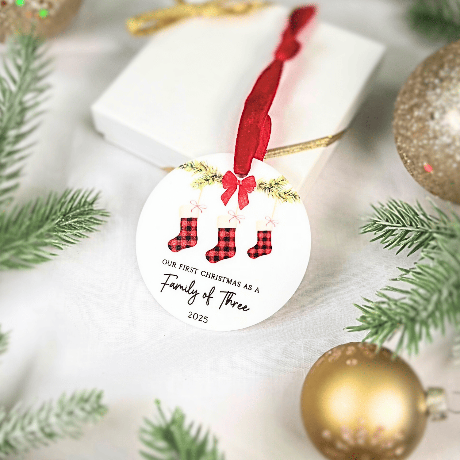 White round acrylic ornament with red plaid stocking design and "Our First Christmas as a Family of Three 2025" text, red ribbon bow attached.