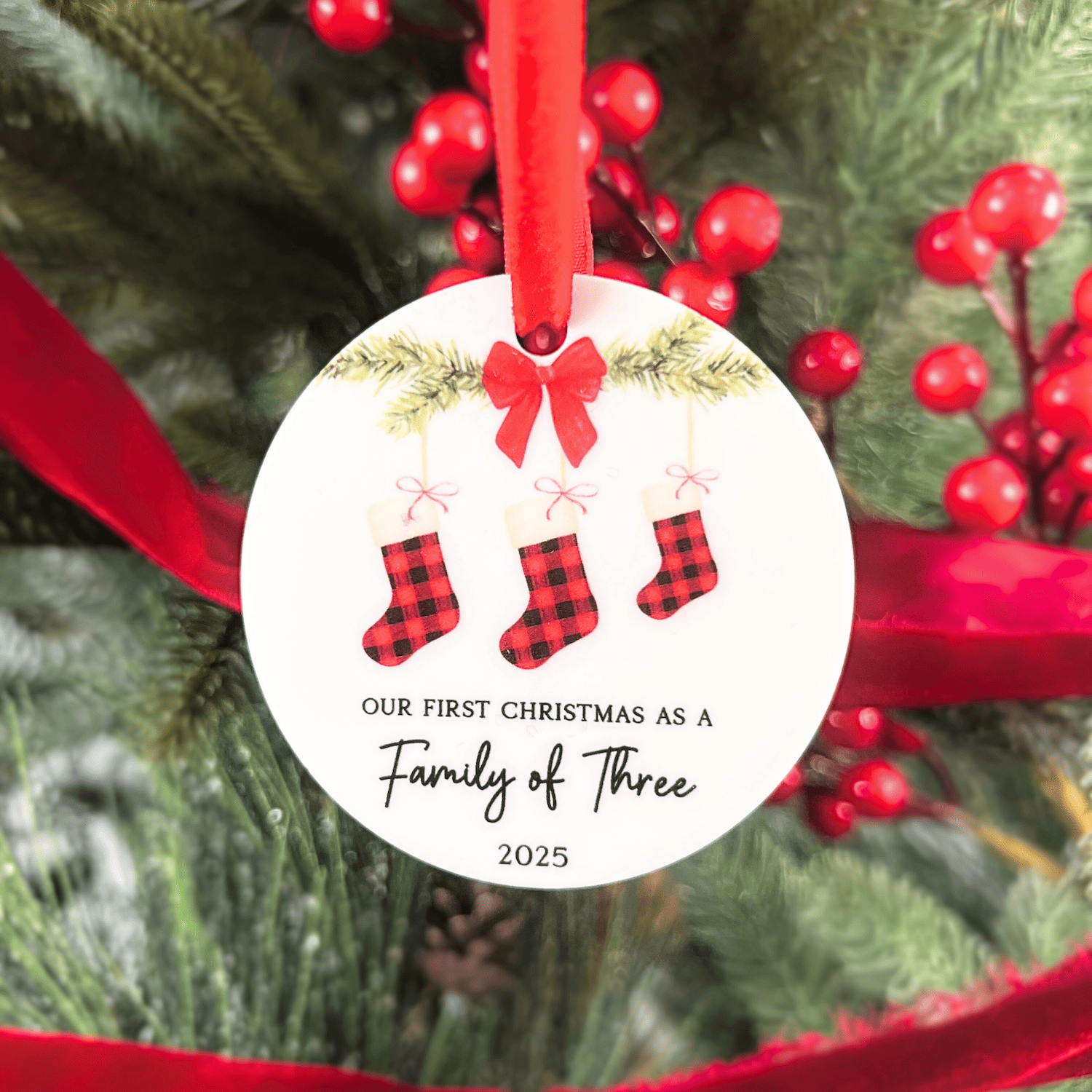 White circular ornament with red ribbon bow, three plaid stockings, and text reading "Our First Christmas as a Family of Three 2025"