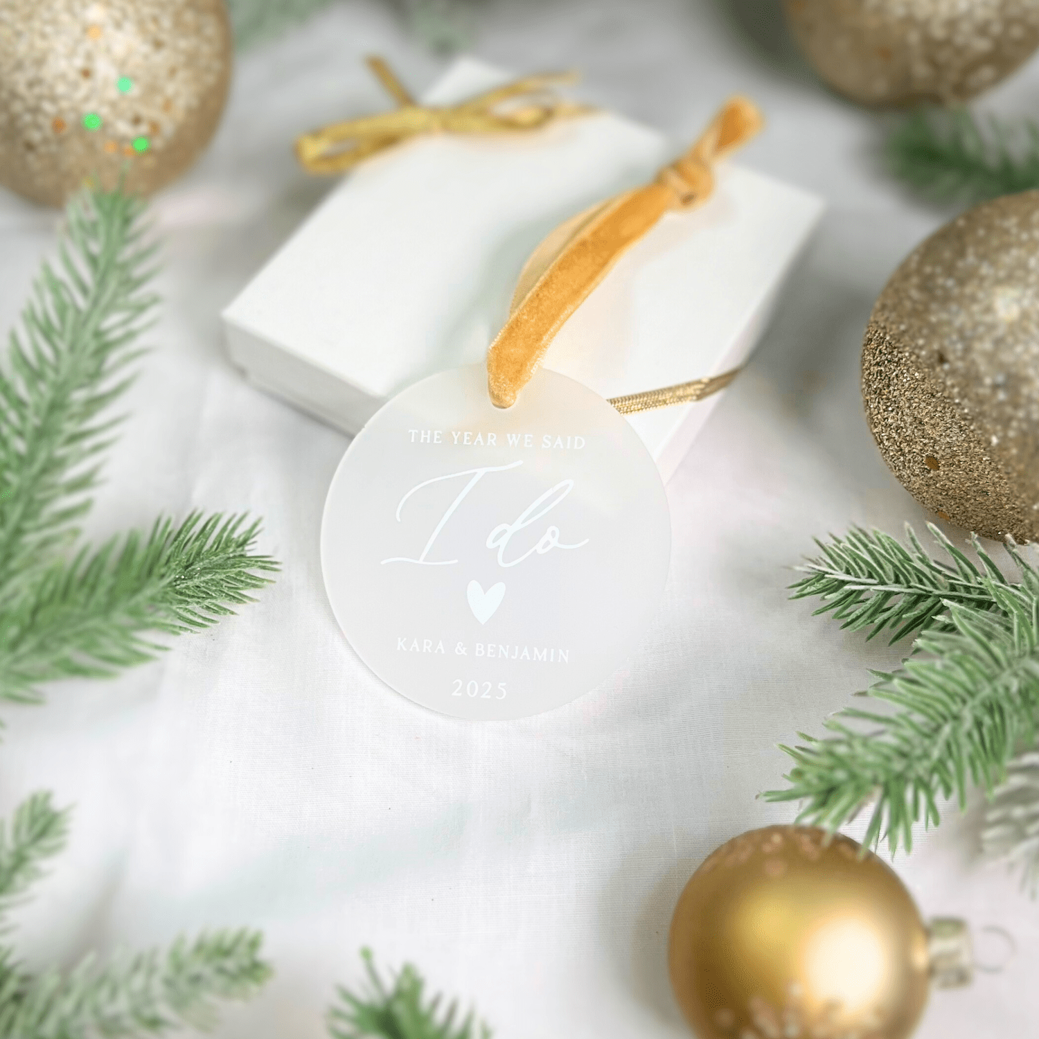 White acrylic ornament with "The Year We Said I Do" engraved text, gold ribbon bow, displayed with evergreen sprigs and gold baubles.