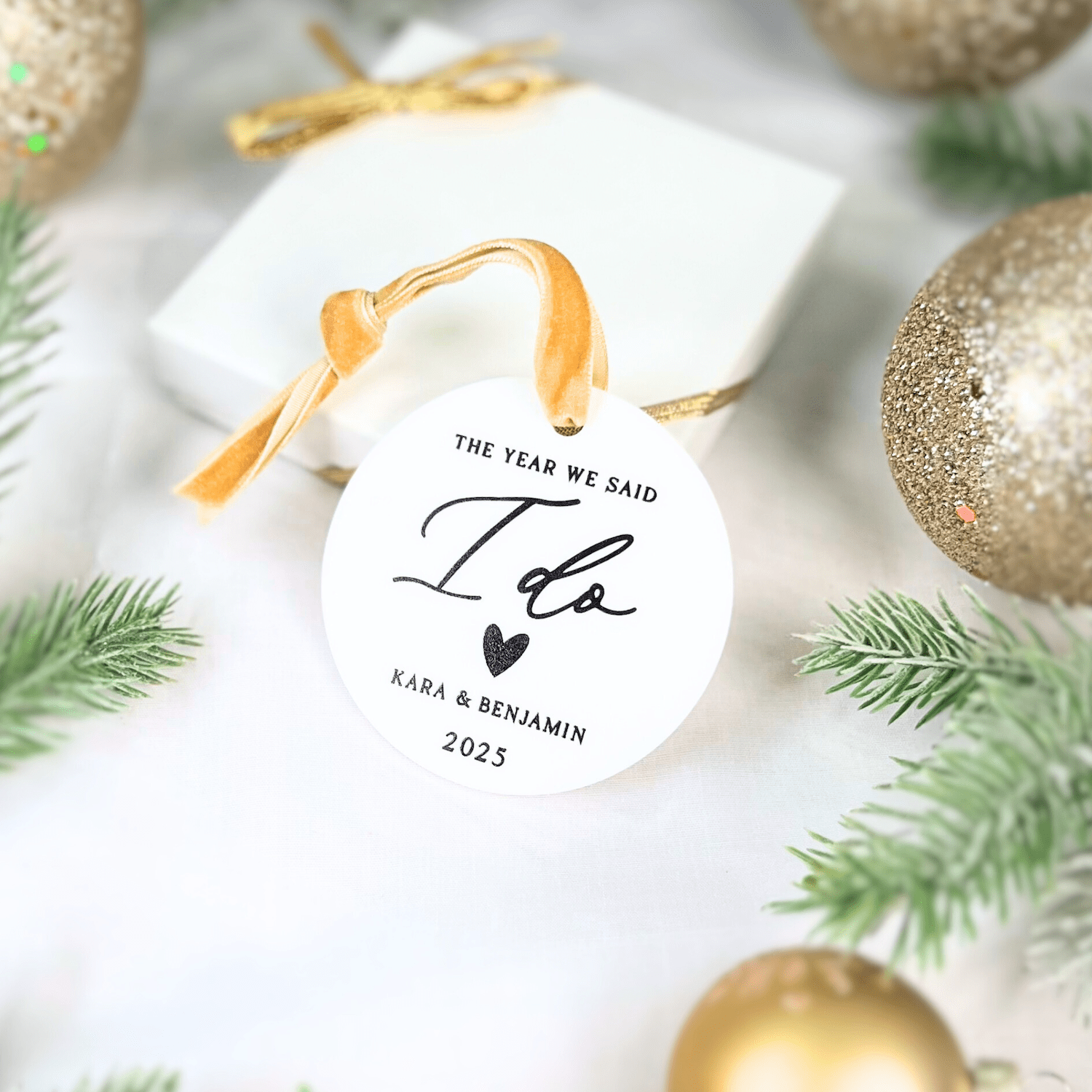 Round white acrylic ornament with gold ribbon, displaying "The Year We Said I Do," couple's names and 2023, surrounded by gold and silver Christmas ornaments and greenery.
