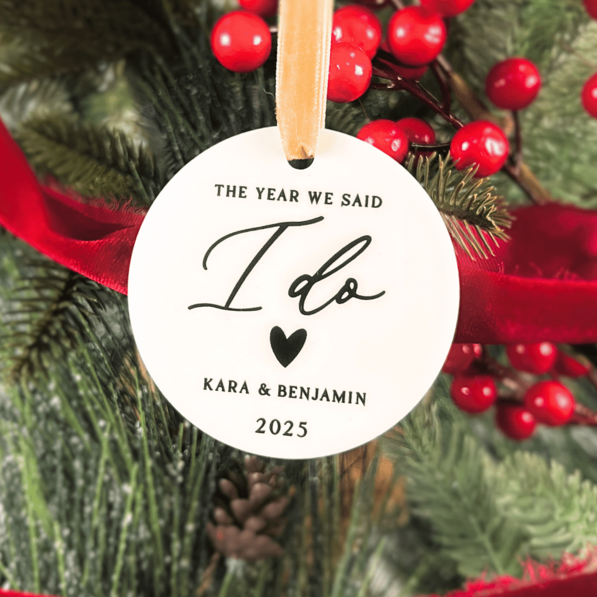 White circular acrylic ornament with "The Year We Said I Do" text, "I do" in script with heart, and "Kara & Benjamin 2025" personalization, wooden string hanger, displayed on festive evergreen and red berry background.
