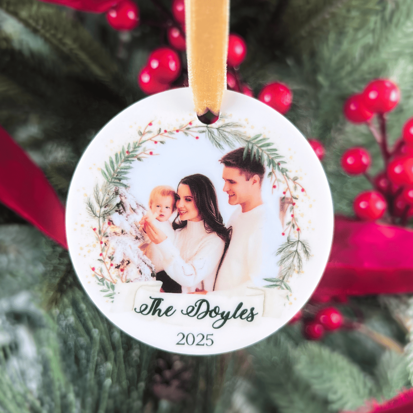 White circular acrylic ornament with family photo, greenery wreath design, personalized name "The Doyles" and year 2025, hanging from tan ribbon.