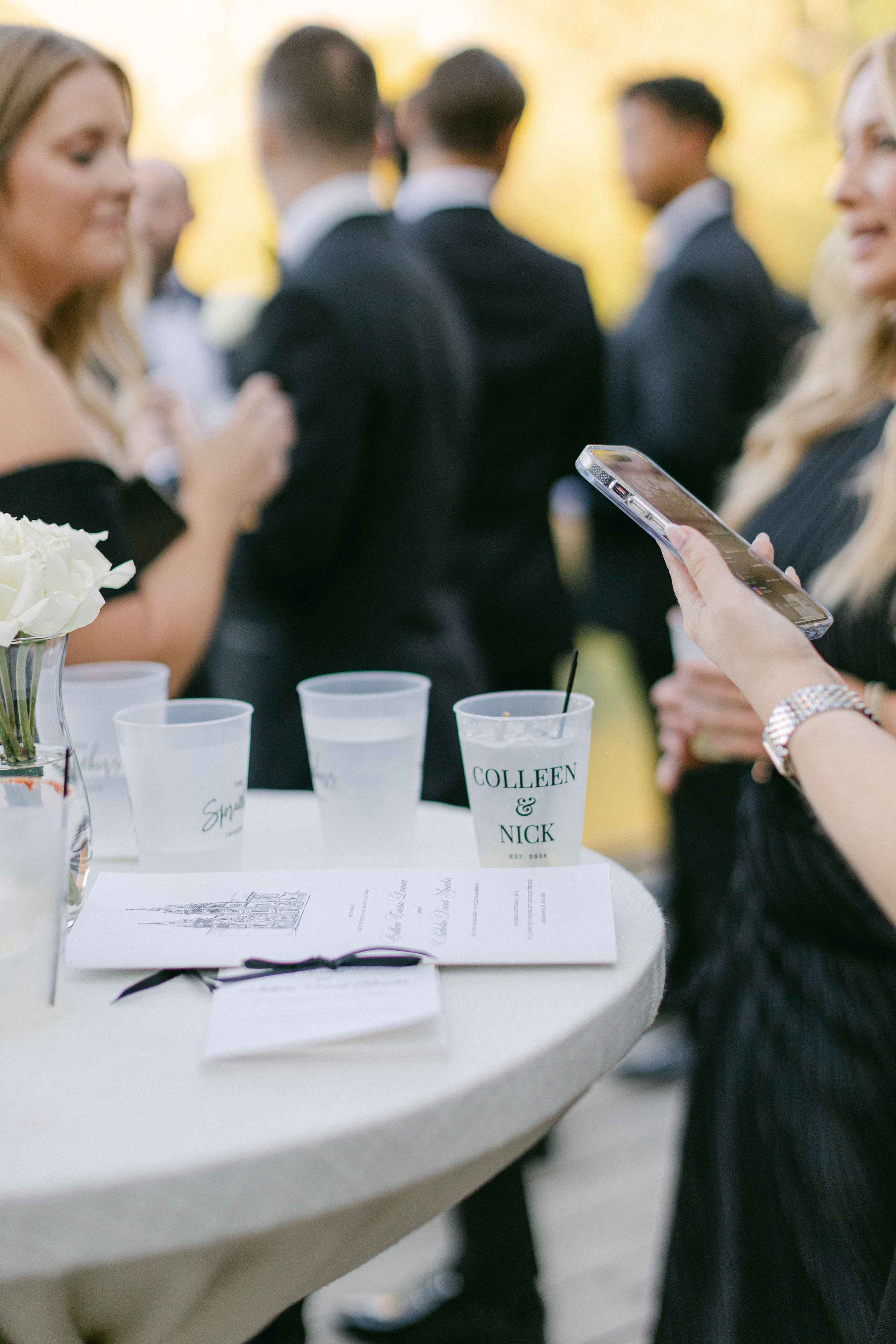 Personalized frosted plastic cups featuring custom names on a white cocktail table at a wedding cocktail hour, with guests and event programs in the background.