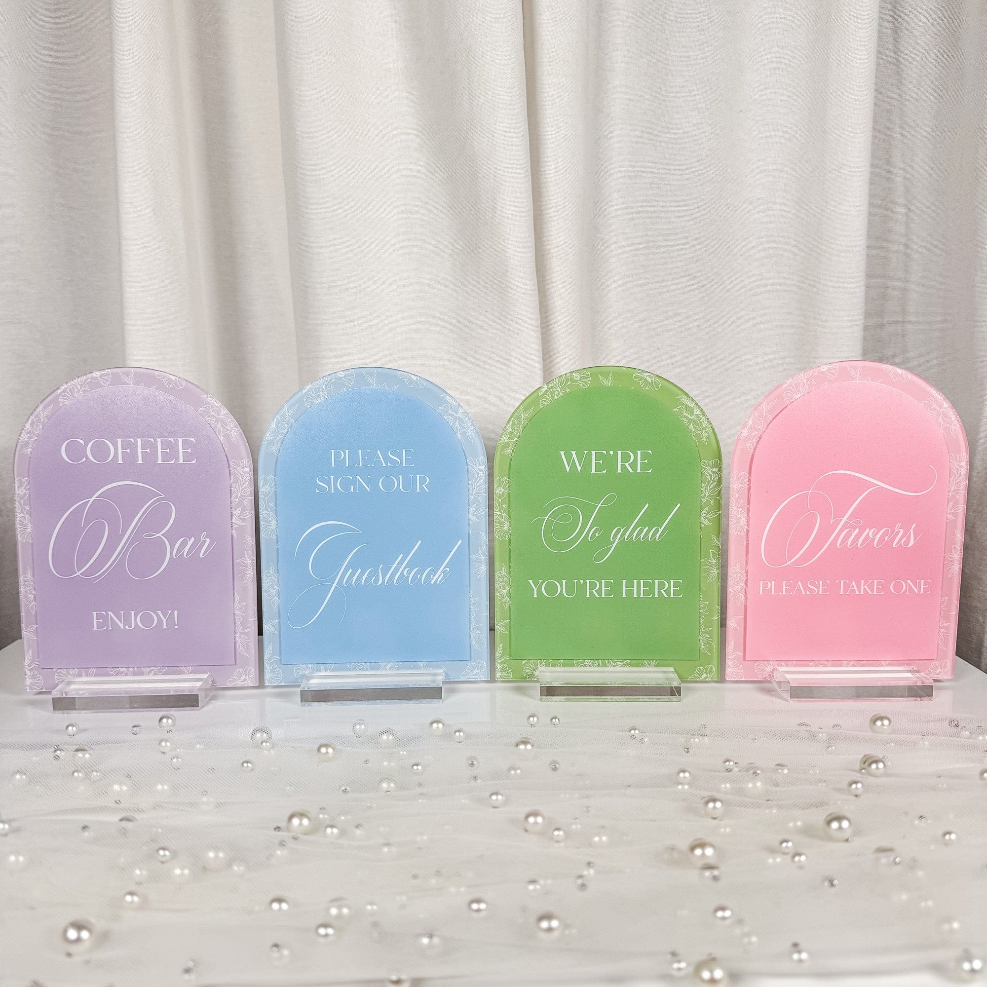 A collection of four arched acrylic event signs in pastel lavender, blue, green, and pink, each featuring a white floral border and standing on a clear base, perfect for guiding guests at a wedding or special occasion.