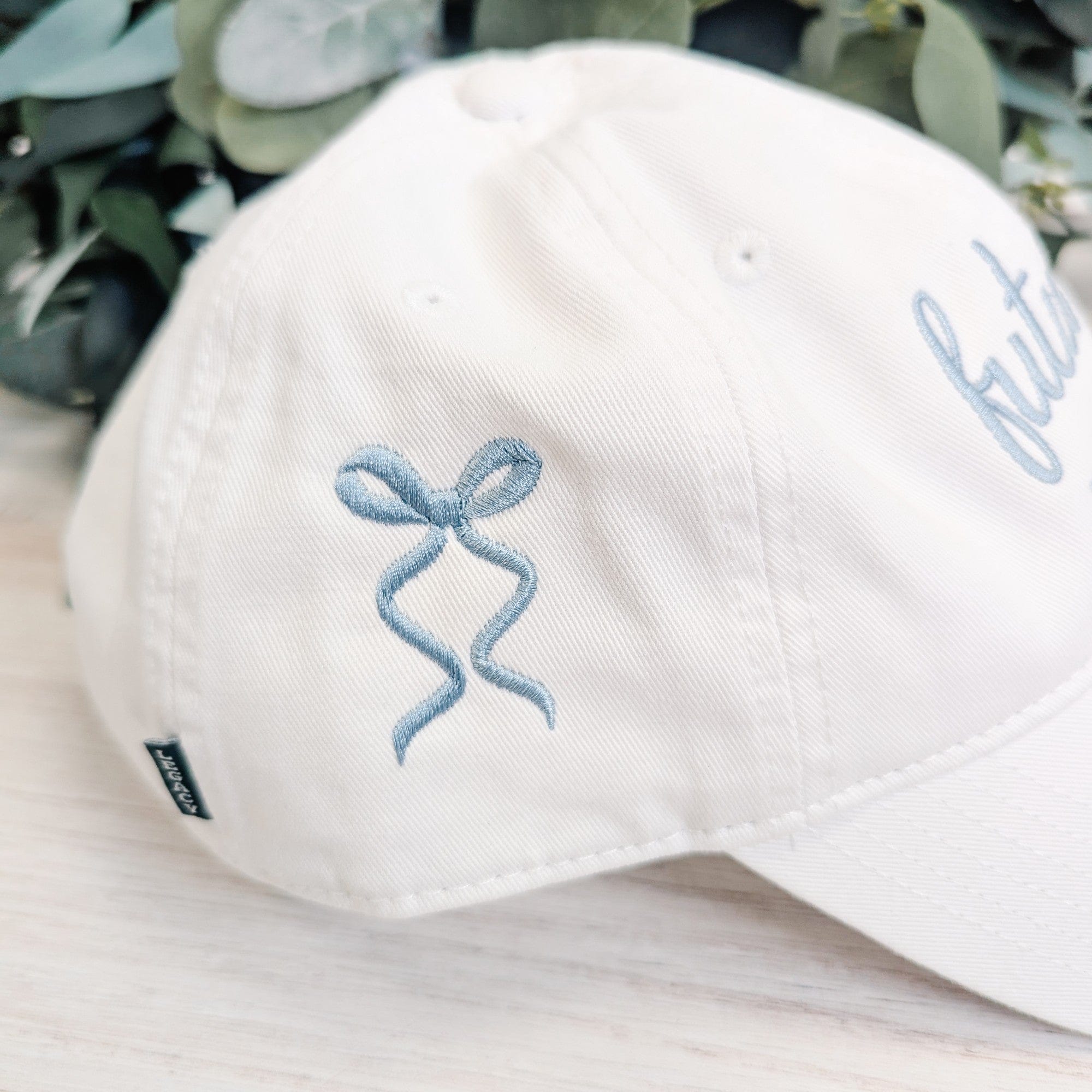 A white cotton baseball cap featuring light blue embroidered ribbon bow detail on the side, designed as a personalized accessory for a bridal event.