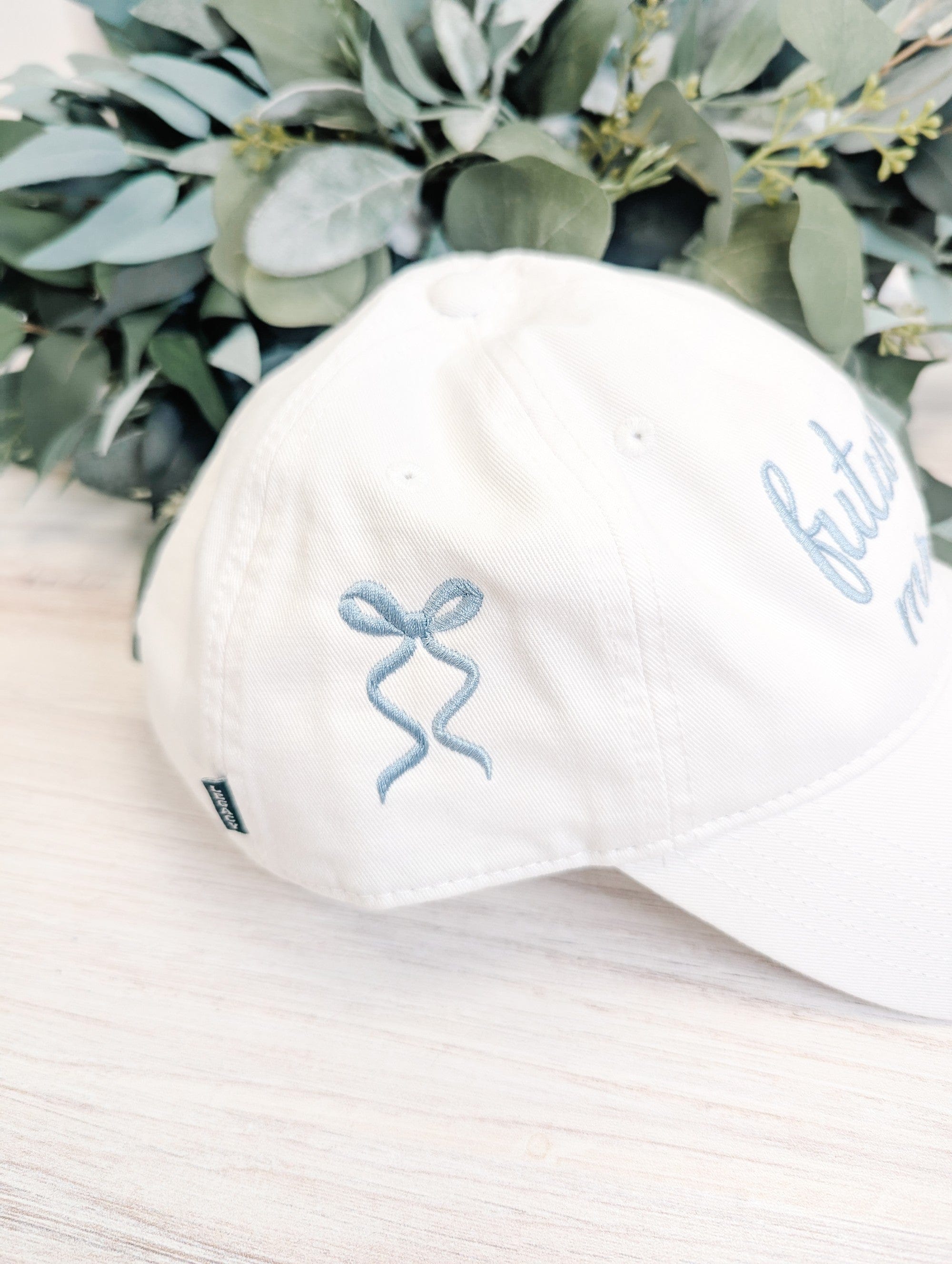 A white cotton baseball cap featuring light blue embroidered bow designs on the side, suitable for a wedding celebration or bridal shower.