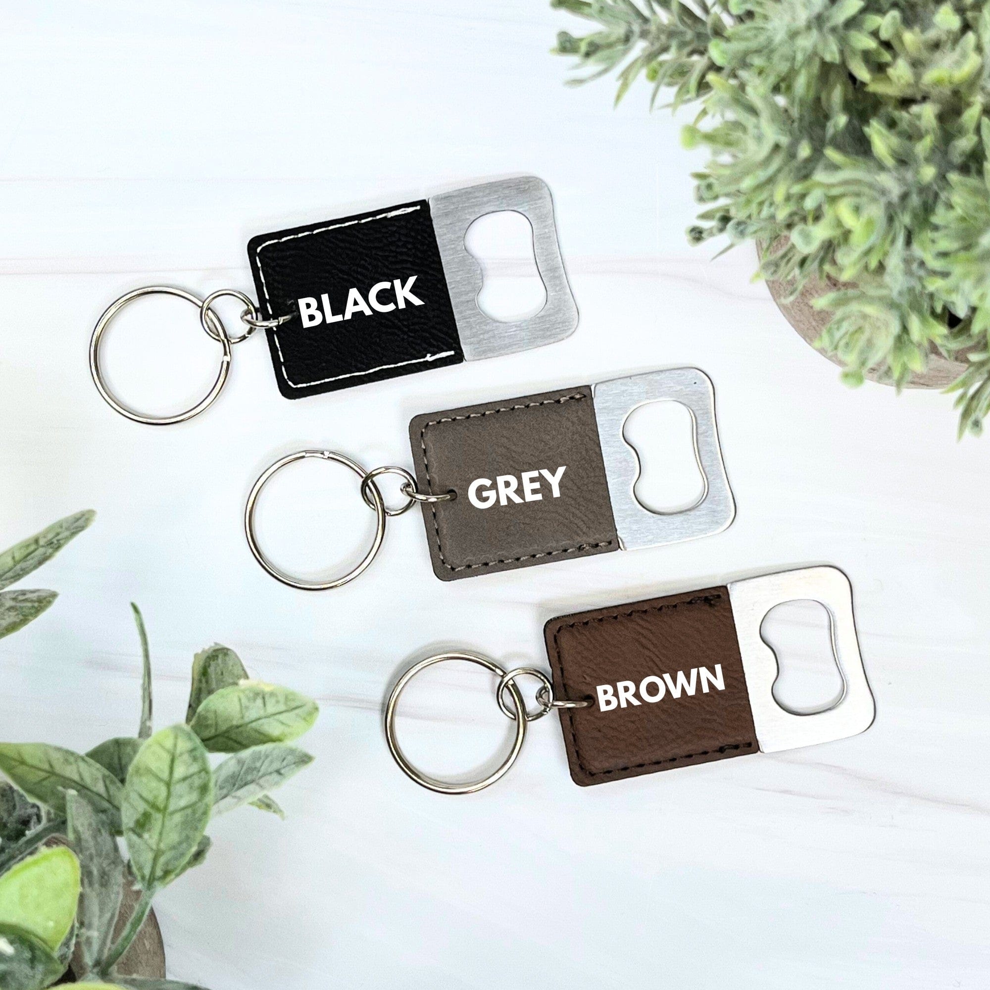 A collection of three bottle opener keychains featuring rectangular faux leather pieces in black, grey, and brown, stitched and attached to a silver-toned metal bottle opener with a keyring, suitable for event favors.