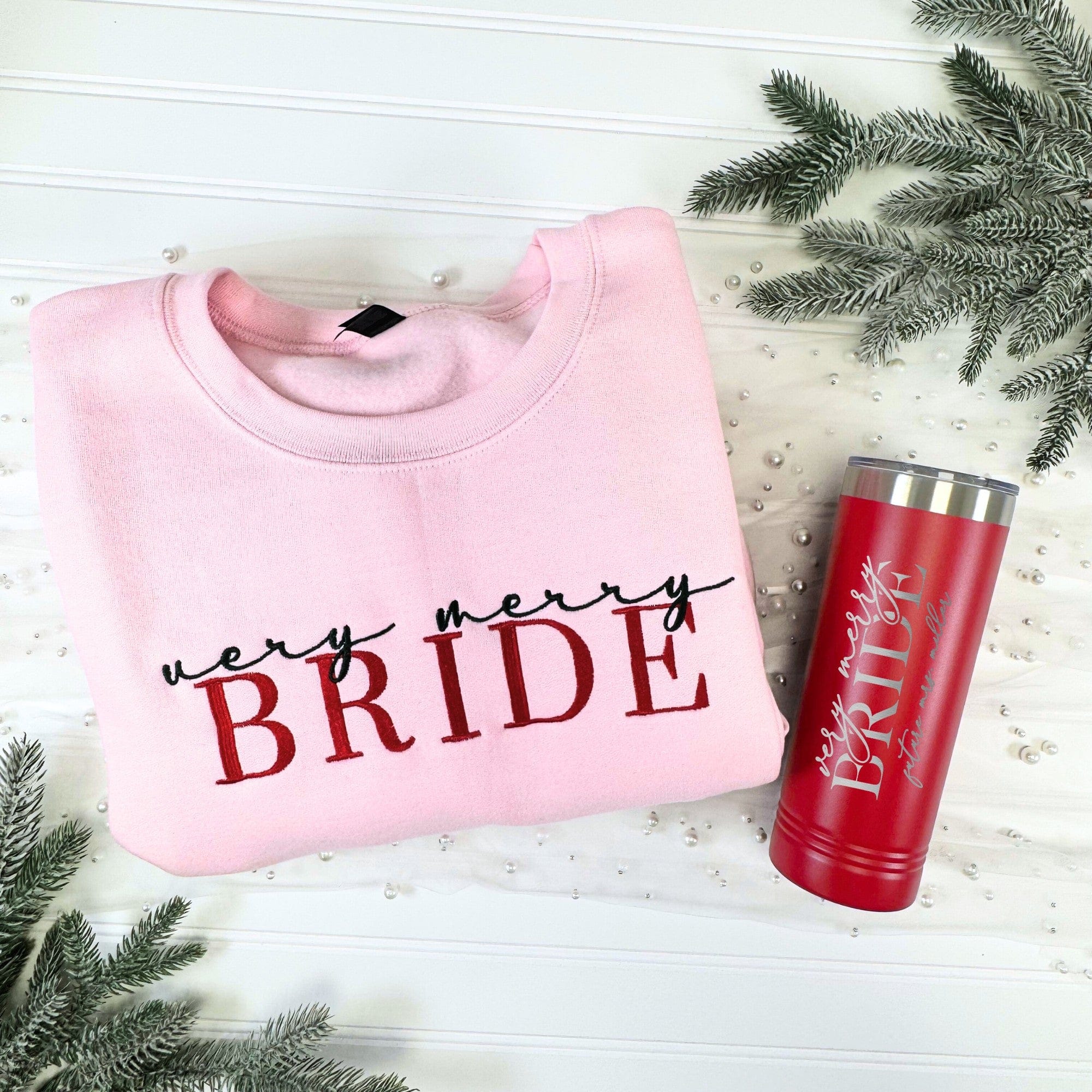 A cozy pink crewneck sweatshirt and a matching red insulated tumbler are showcased, both personalized for a bridal event, surrounded by festive frosted pine branches and shimmering pearls.