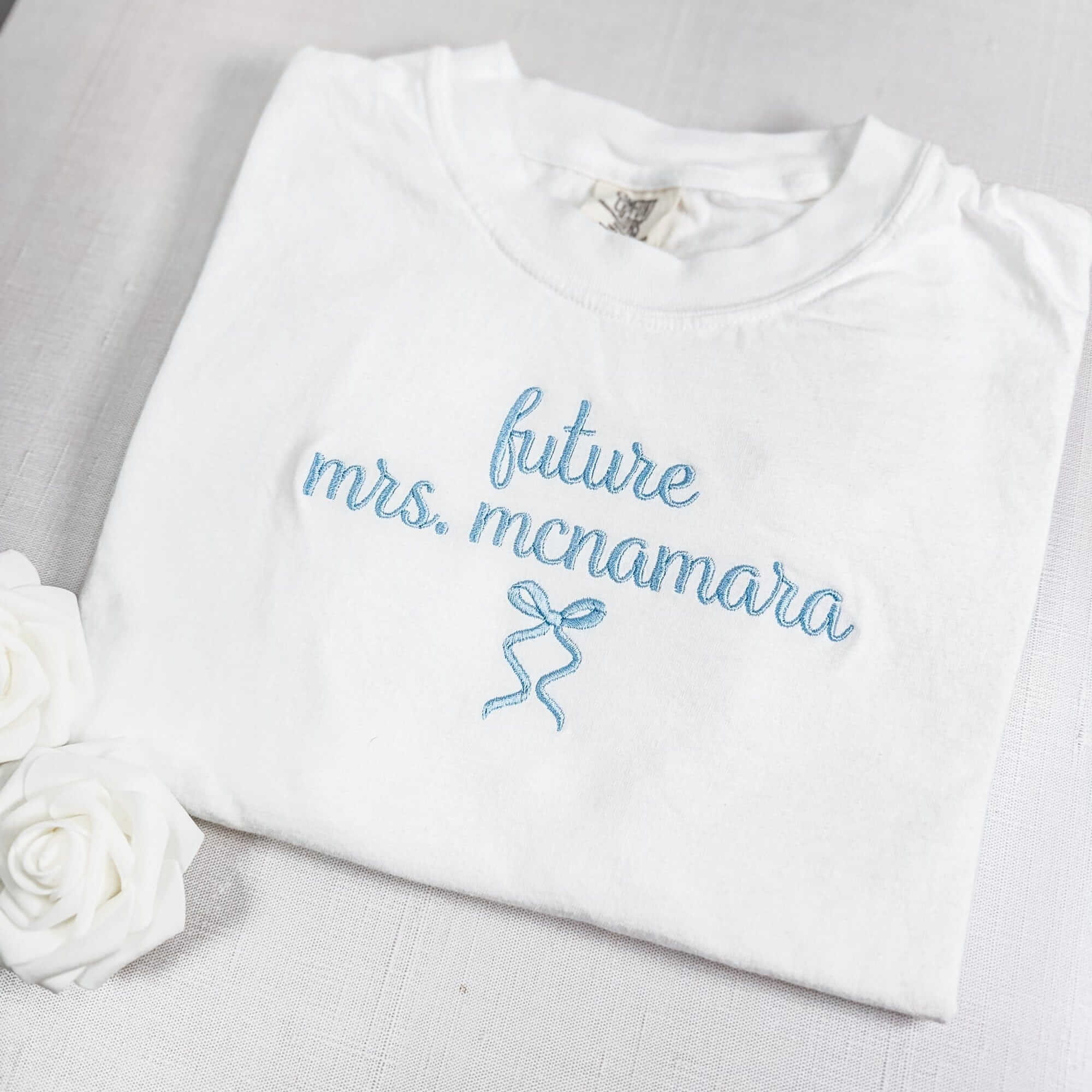 A folded white cotton t-shirt features light blue embroidered text with a delicate bow design, ideal for a bridal shower or pre-wedding event.