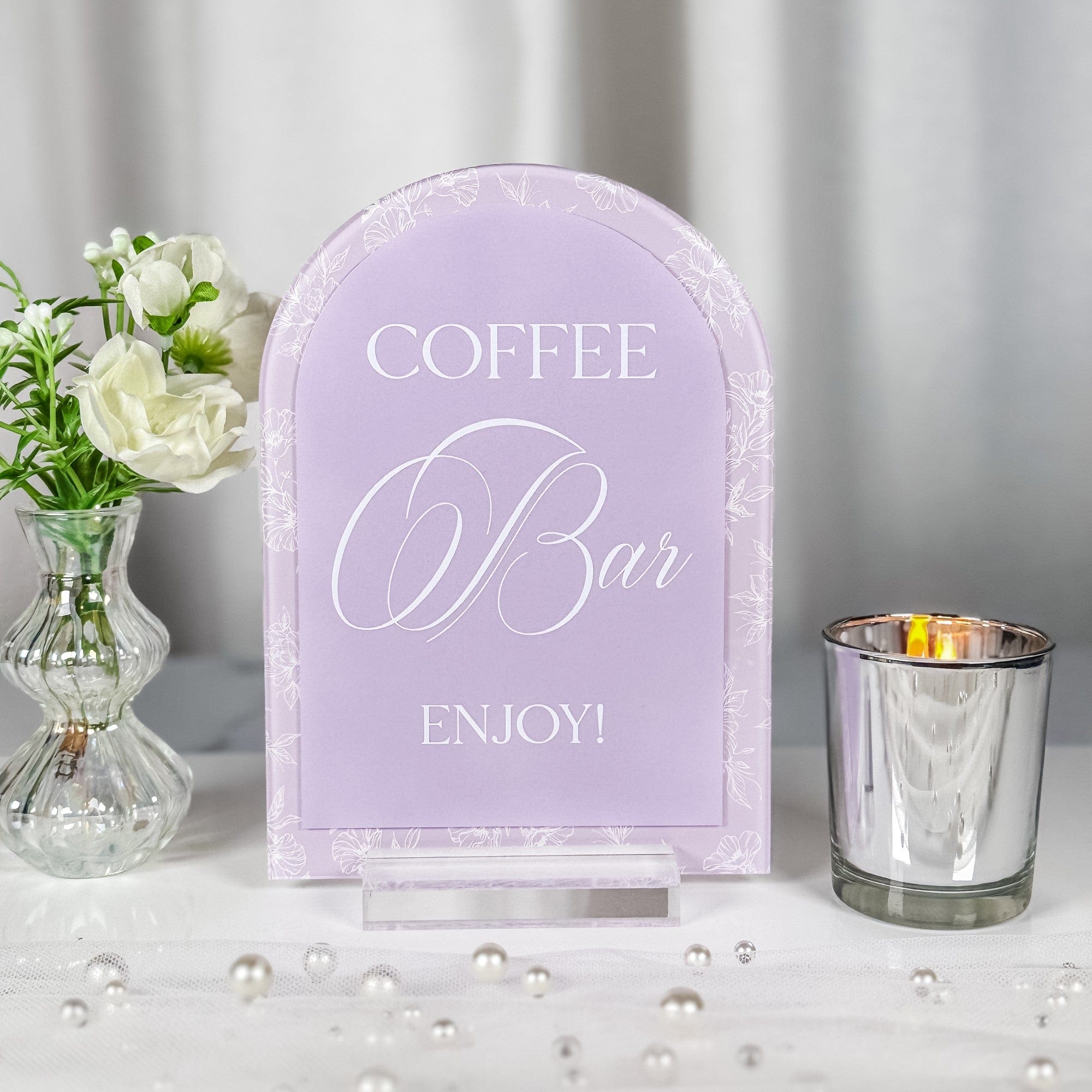 An arched lavender acrylic coffee bar sign with white floral outline detailing, displayed on a clear stand, perfect for a wedding or special event.