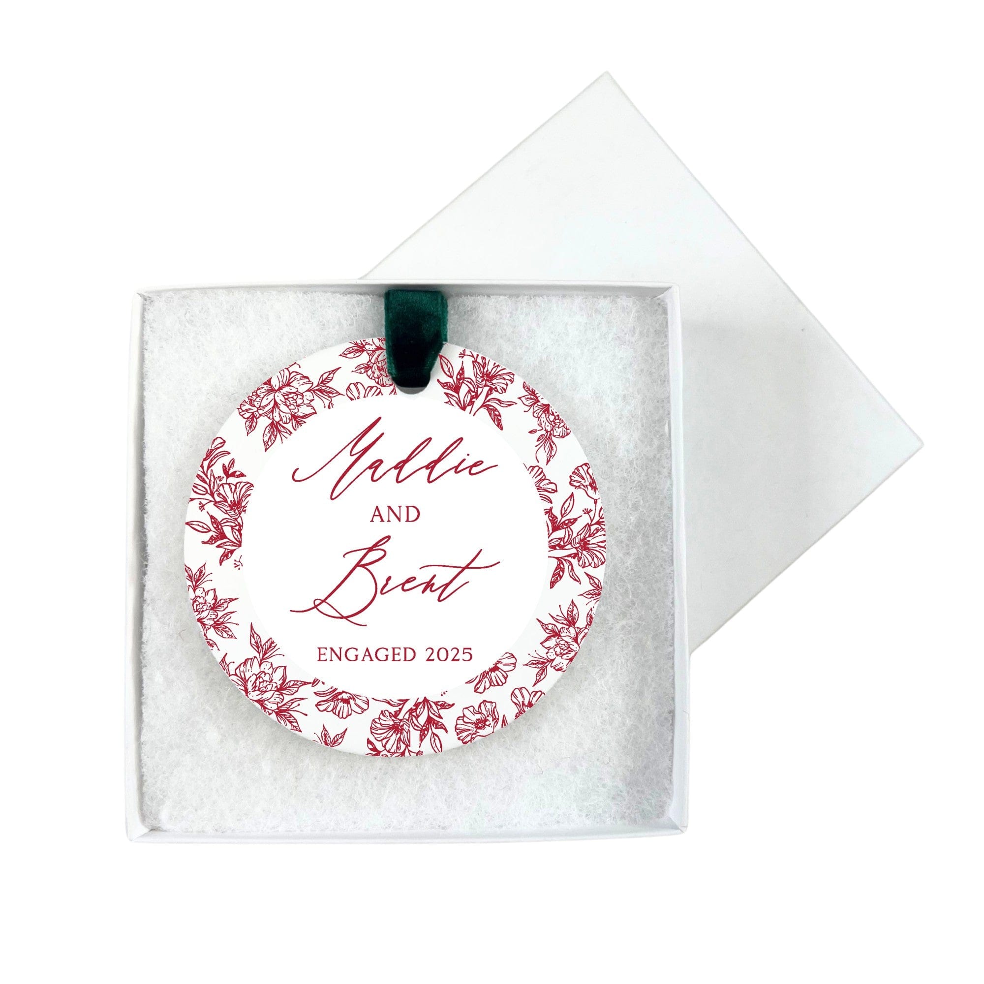 A circular white engagement ornament featuring a red floral border design, suspended by a green velvet ribbon, and presented in an open white gift box.