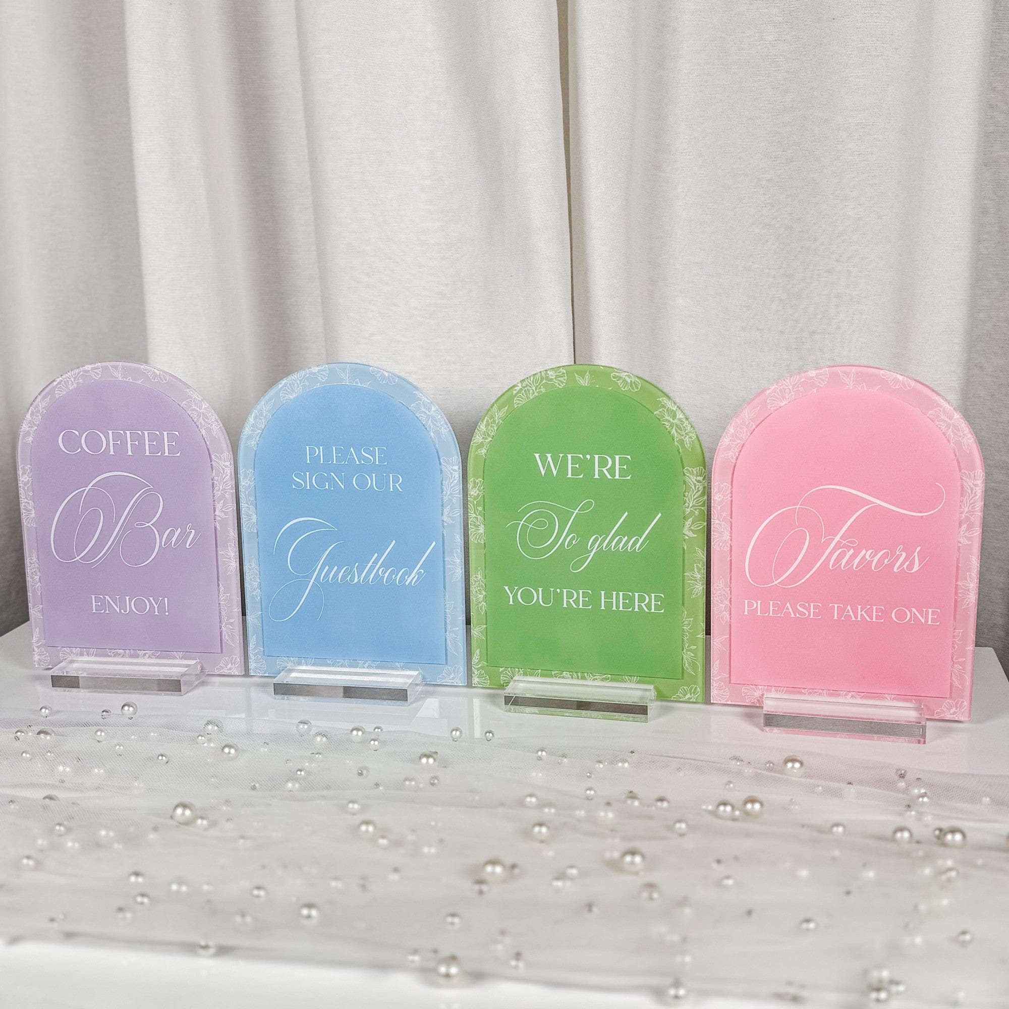 Four arch-shaped acrylic event signs in pastel lavender, light blue, lime green, and pink, each featuring a delicate white floral border and a clear acrylic stand, designed to guide guests at a wedding or event.