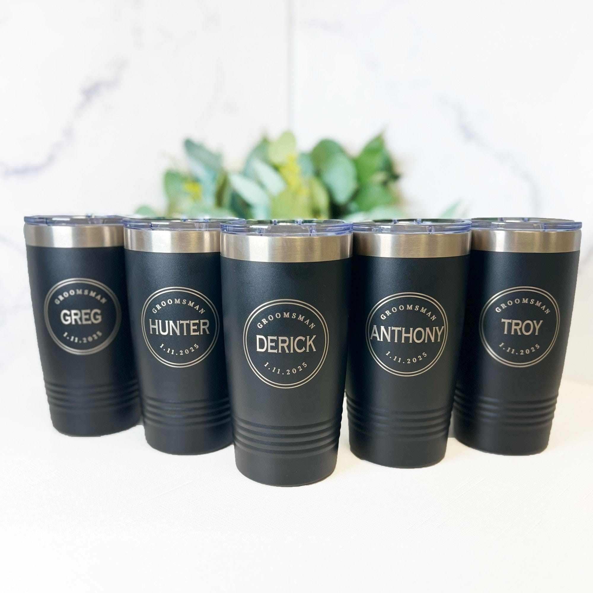 Five personalized black insulated tumblers with silver tops and clear lids, each featuring a circular engraved design, perfect for a wedding party.