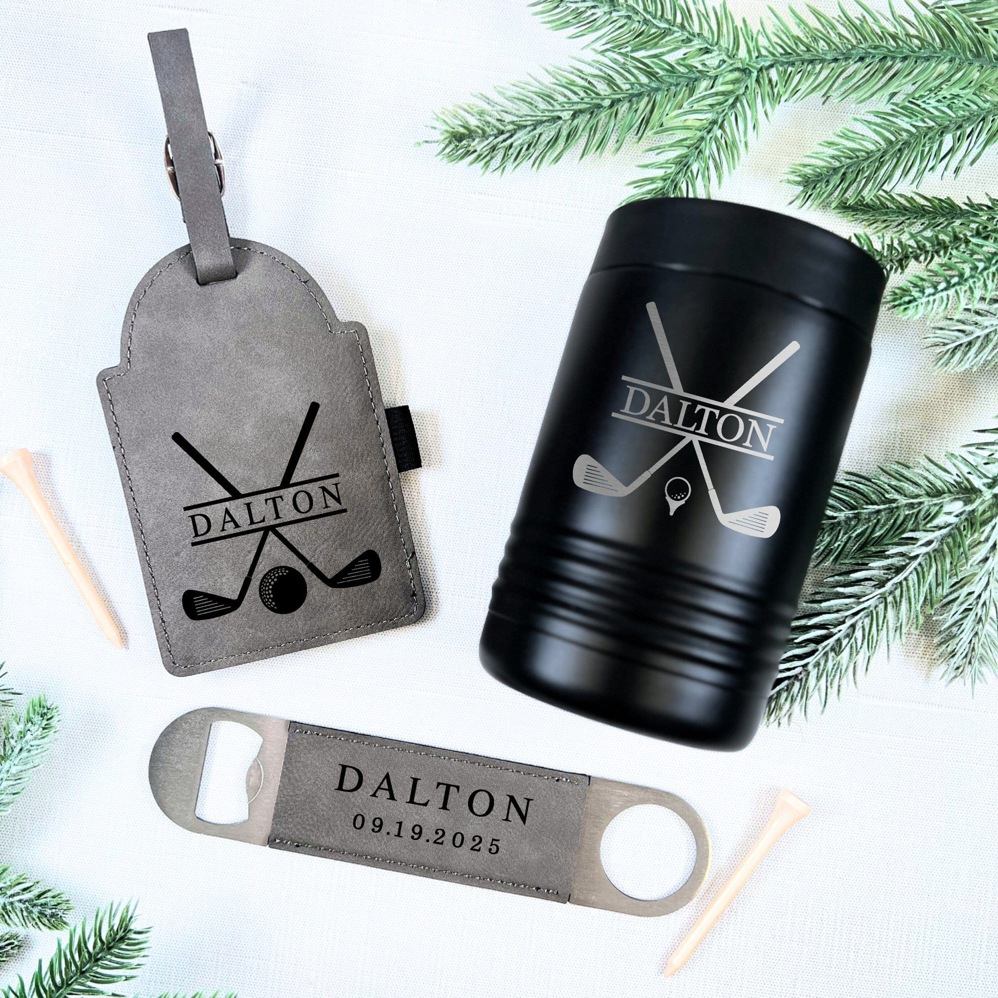 A personalized groomsmen golf gift set featuring a grey faux leather luggage tag, a black insulated metal can cooler, and a silver and grey bottle opener, all designed with a golf club and ball motif, displayed on a white background with green pine accents.