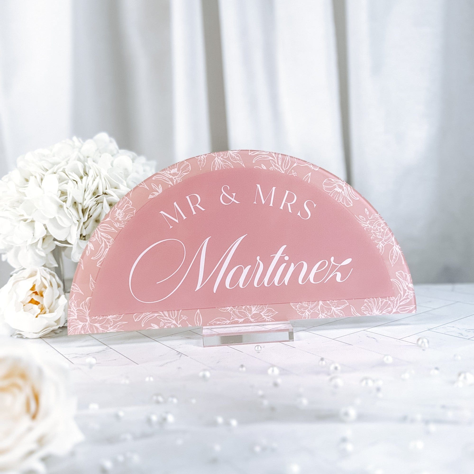 A dusky pink acrylic half-arch wedding sign with a white floral border and white script text, displayed on a clear stand for a wedding reception.