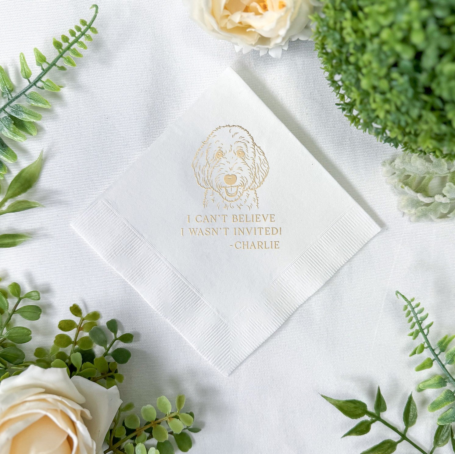 A white folded paper napkin featuring a custom gold foil pet illustration, intended for personalized wedding or event decor.