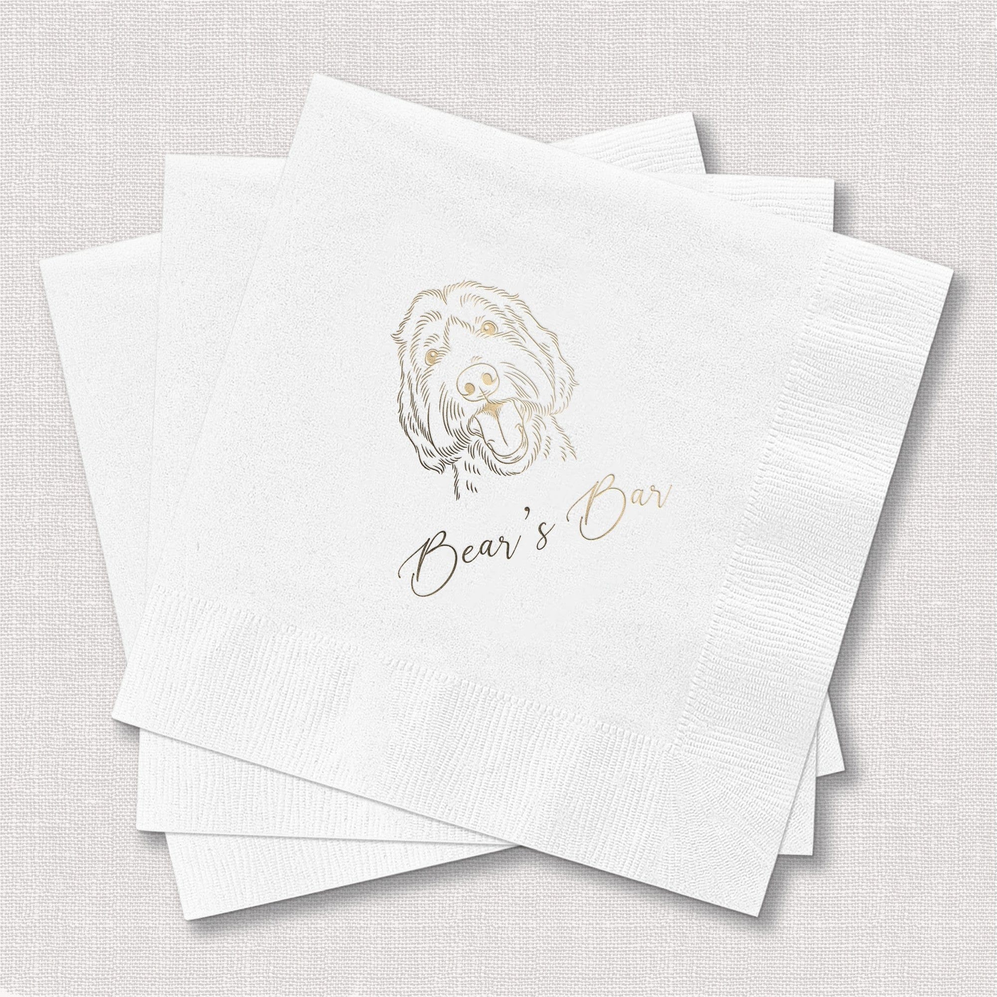 A stack of white paper cocktail napkins featuring a custom gold foil illustration of a dog and elegant script, ideal for a personalized wedding or event bar.