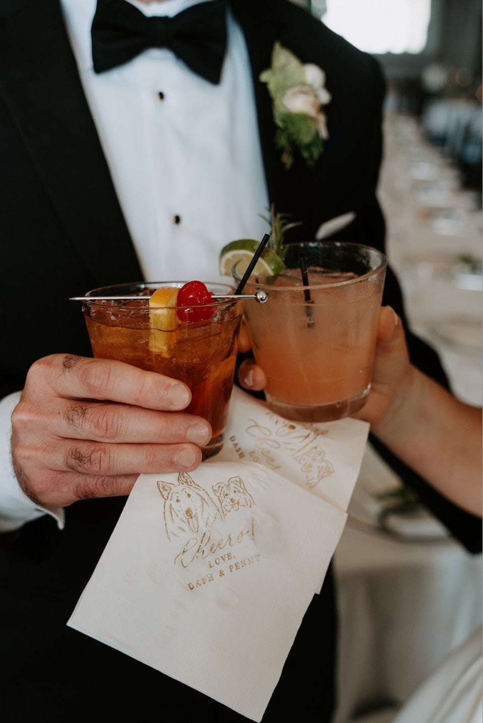 A person in a tuxedo holds two cocktails over custom white paper napkins featuring gold foil dog illustrations and personalized text, at a wedding reception.