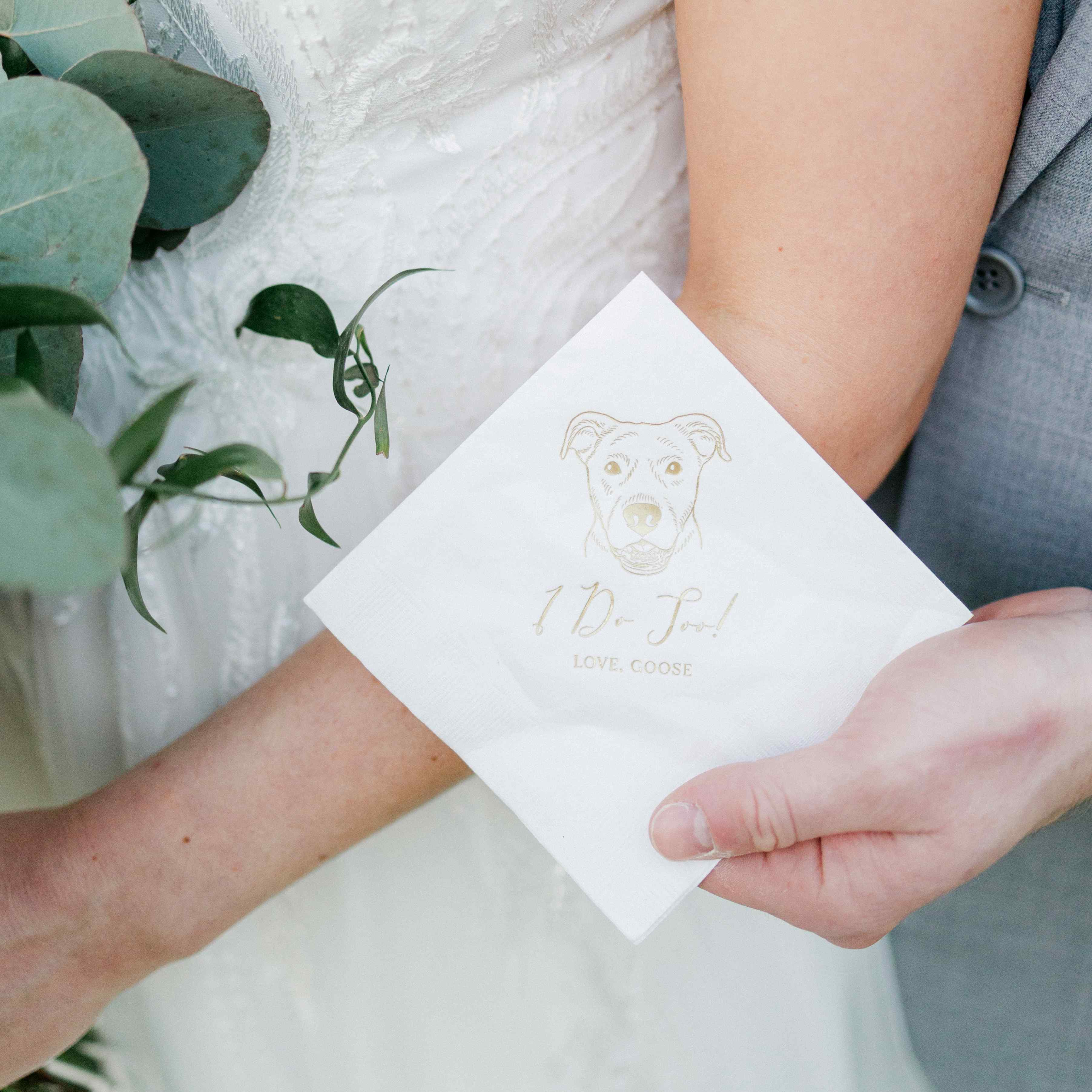 Close-up of a white paper wedding napkin featuring a gold foil custom dog illustration and 'I Do Too!' text, held by hands in a bridal setting.
