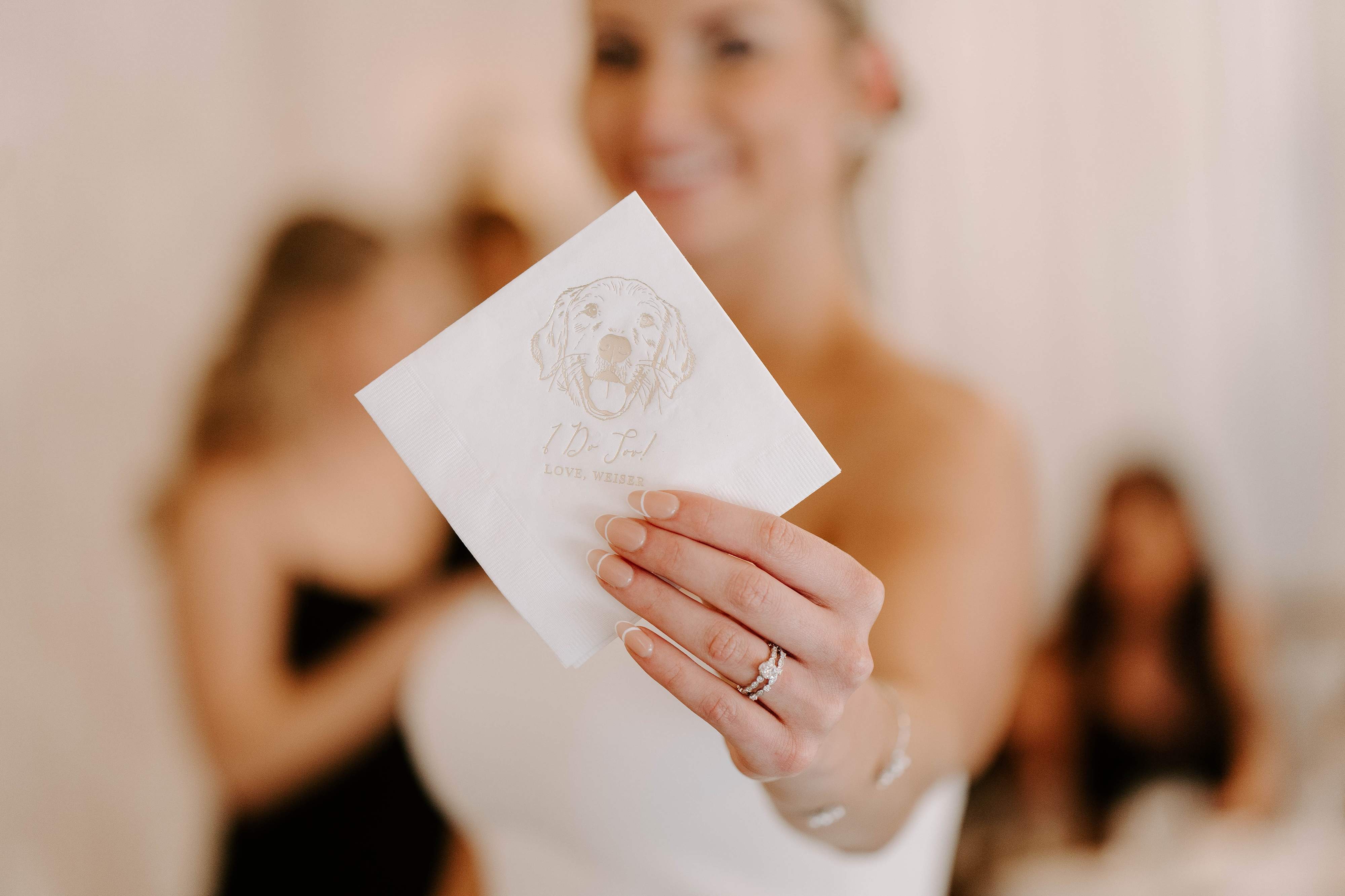 A close-up of a bride's hand holding a white paper wedding napkin featuring a gold foil dog illustration and custom 'I Do Too!' text, perfect for pet-inclusive wedding celebrations.