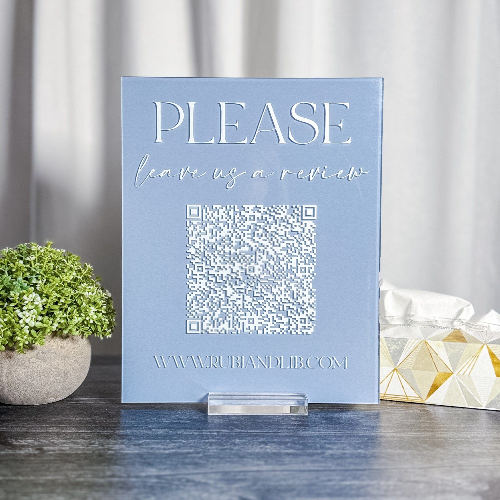 A light blue acrylic review sign featuring white raised lettering and a white QR code, standing on a clear acrylic block, designed to encourage customer feedback at a wedding or event.