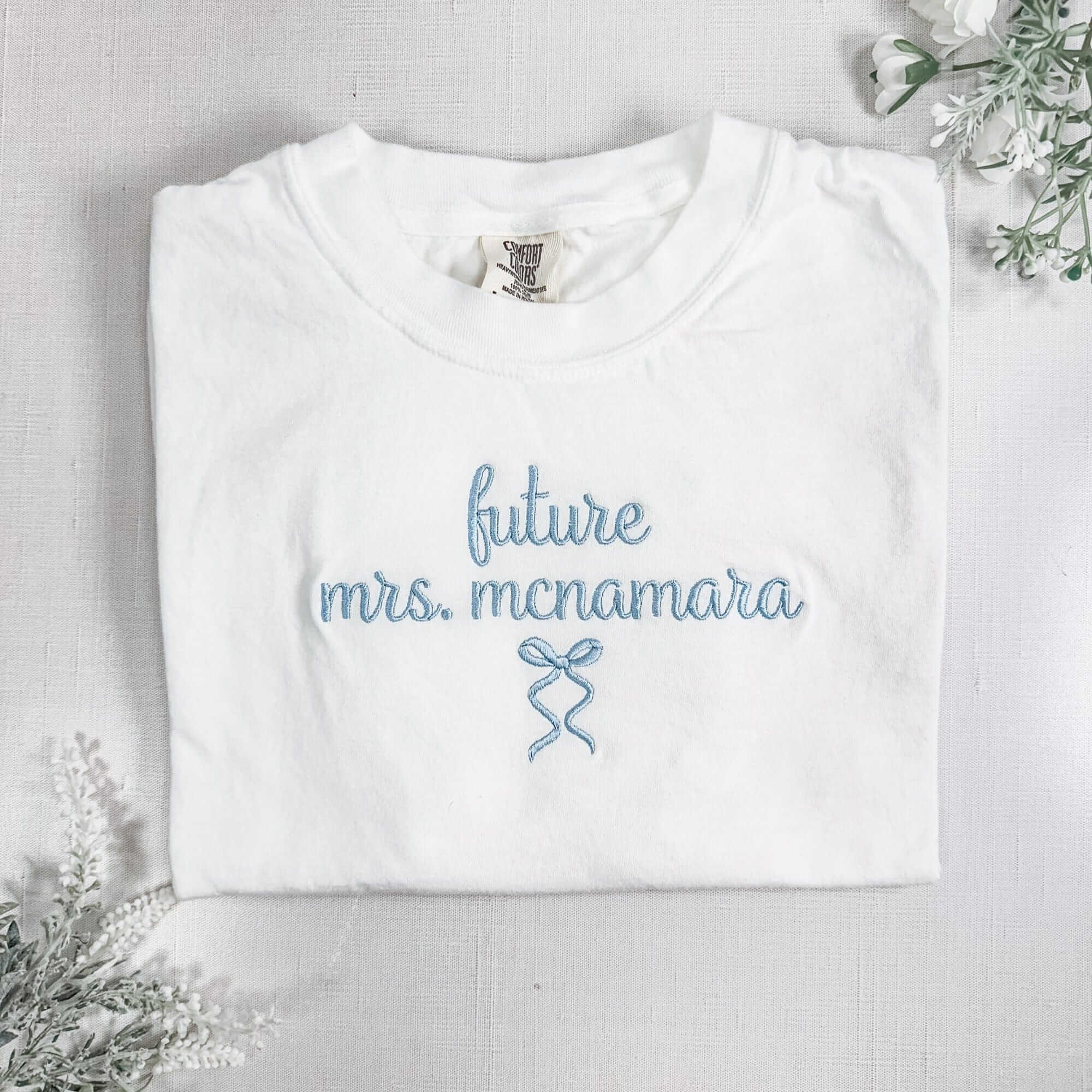 A folded white cotton t-shirt features light blue embroidered text and a delicate bow design, suitable for bridal events.
