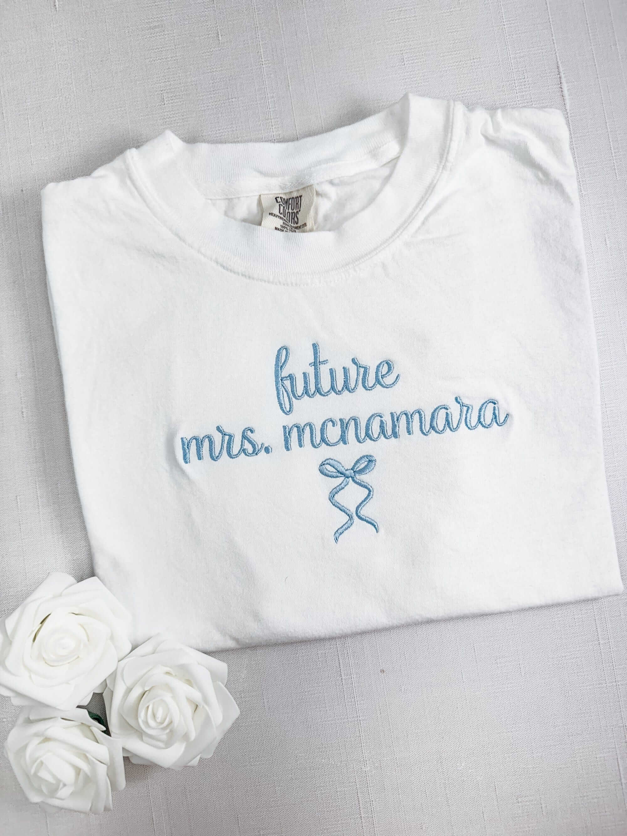 A white cotton t-shirt featuring light blue embroidered script text and a small bow design, ideal for a bridal event.
