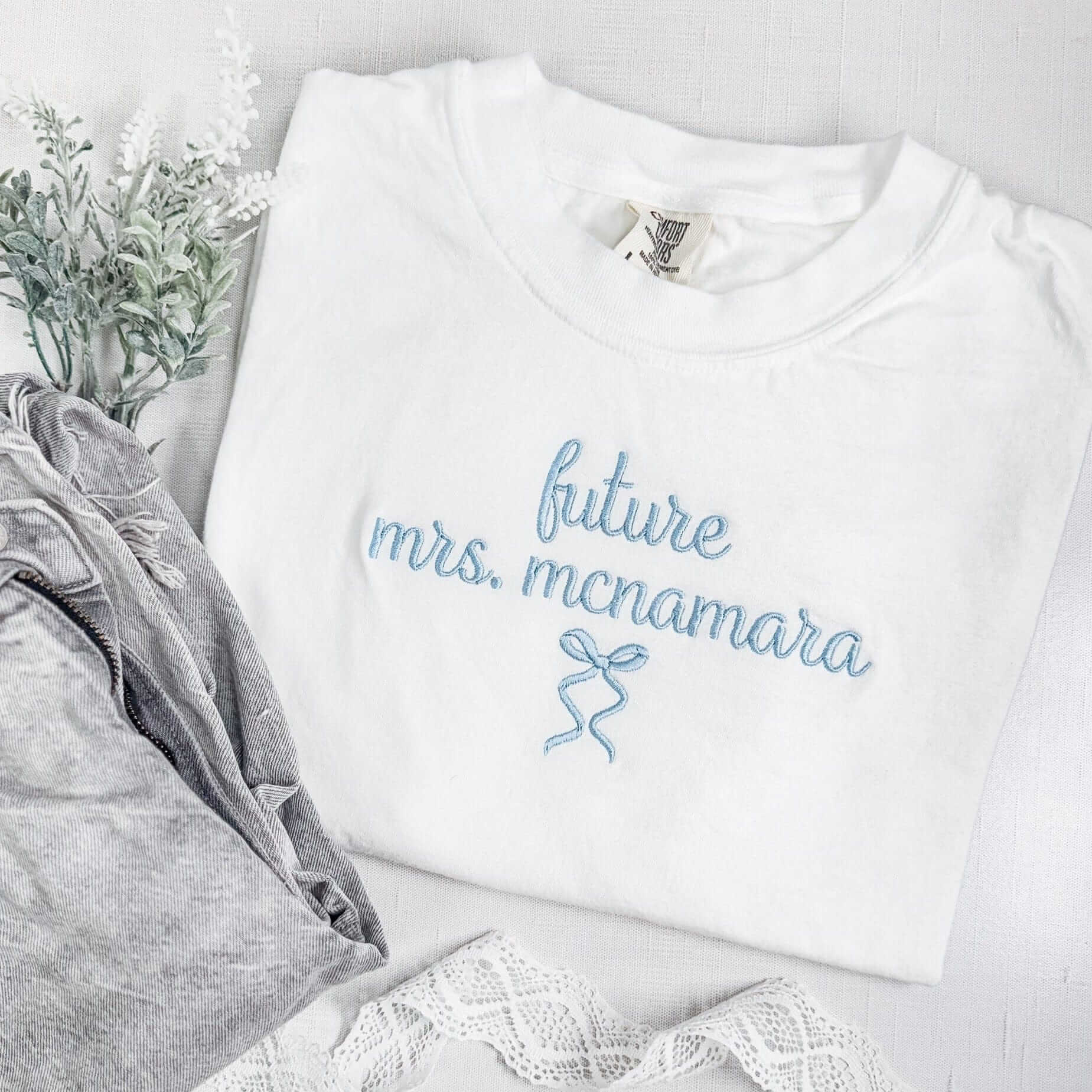 A white short-sleeve t-shirt featuring elegant light blue embroidery with a future bridal status design and a small decorative bow, suitable for wedding-related celebrations.