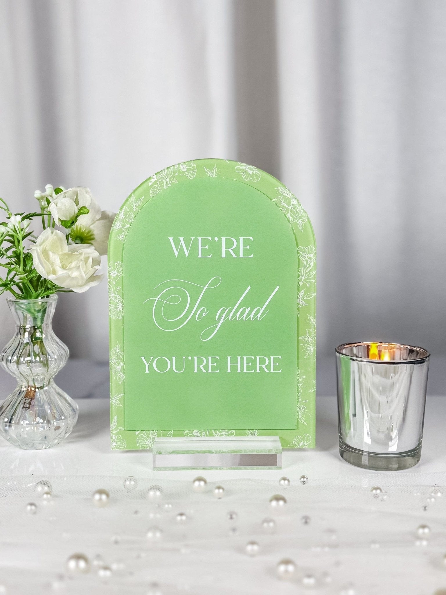 A pistachio green arched acrylic event sign featuring white floral border details and white text, displayed on a clear acrylic stand amidst white flowers and pearls.
