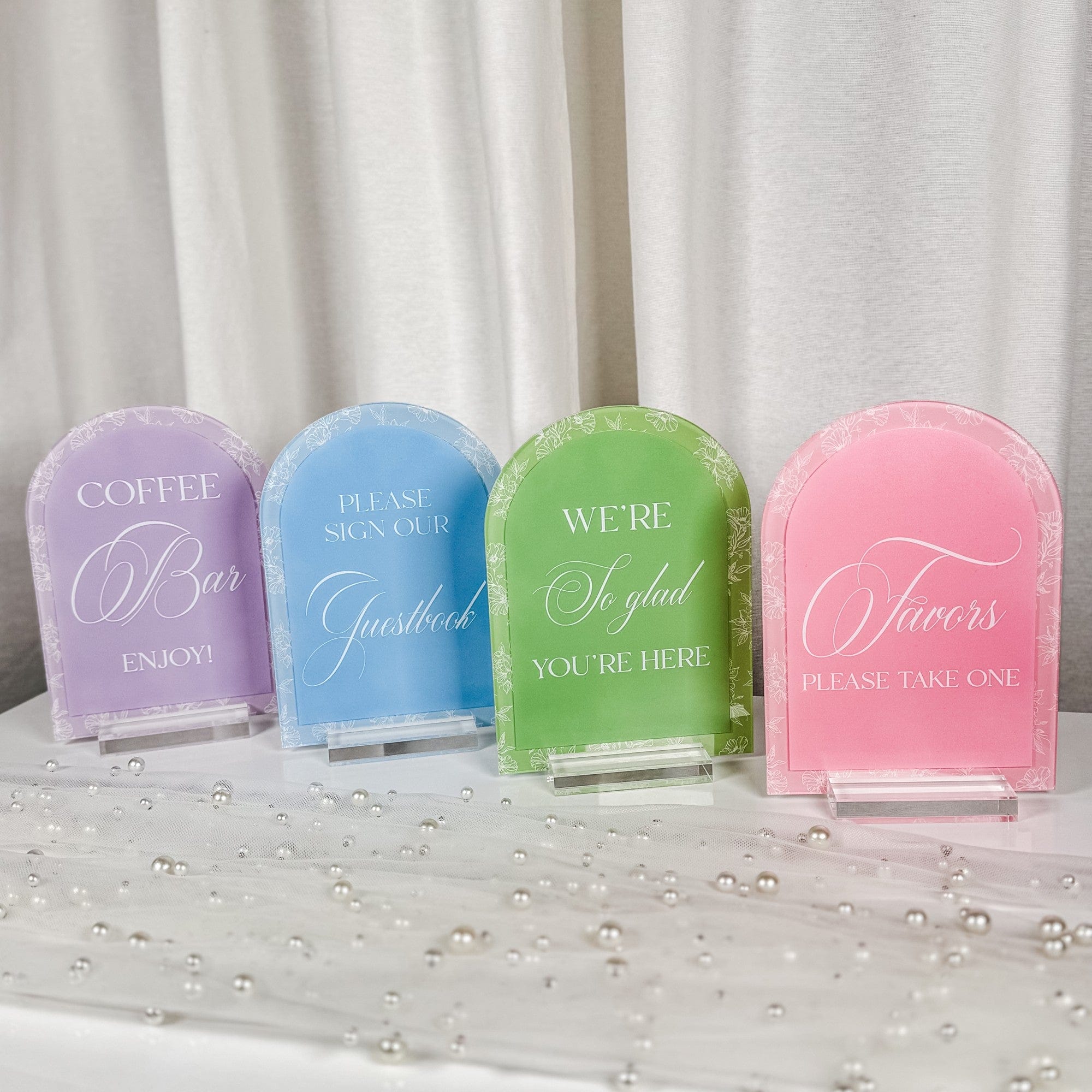 A set of four arched pastel-colored acrylic signs featuring white text and delicate floral outline borders, displayed on clear acrylic stands, perfect for wedding or event table decor.