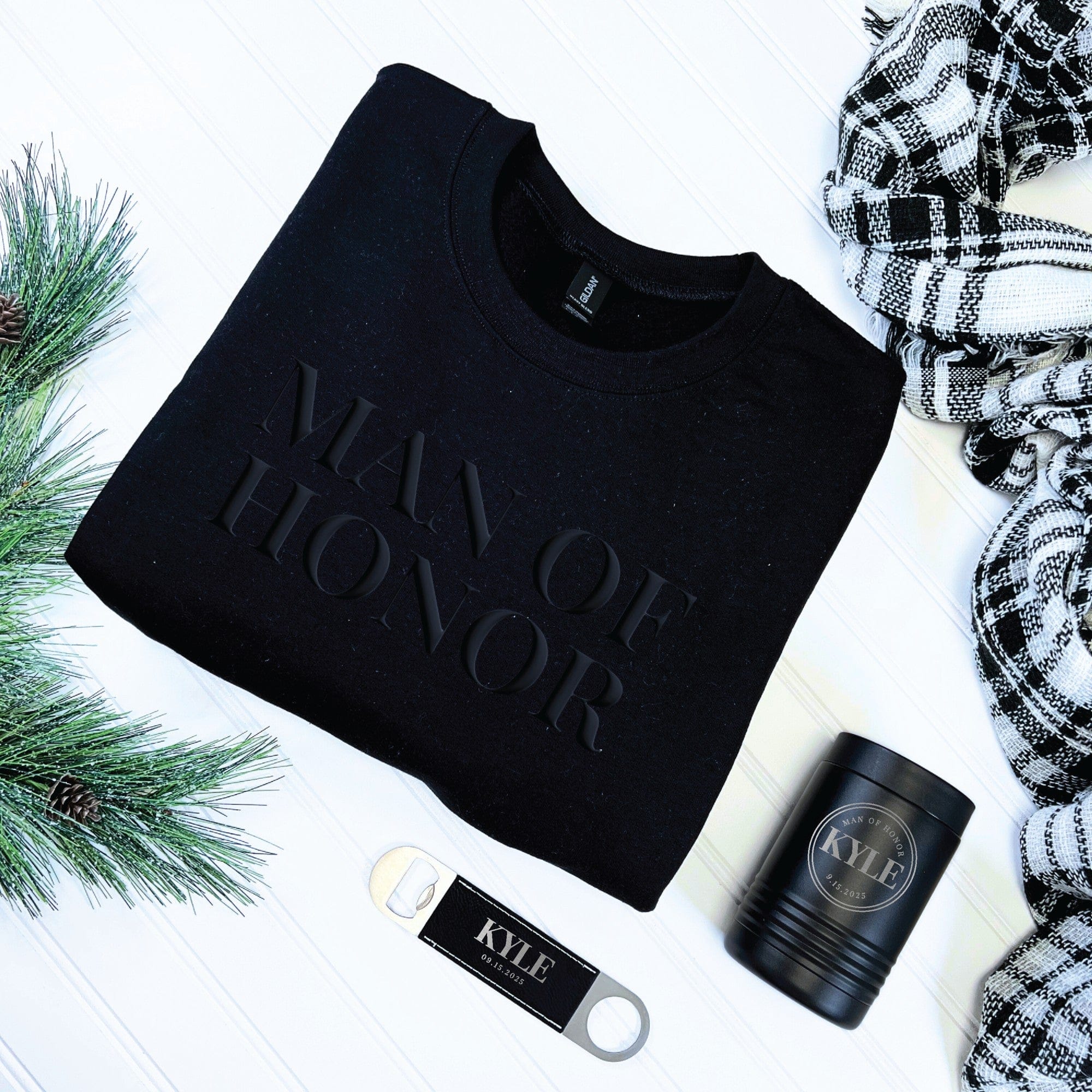 A black long-sleeved sweatshirt with a raised text design is displayed alongside a black insulated can cooler with an engraved design and a silver bottle opener with a black leatherette grip, all part of a personalized wedding party gift set.