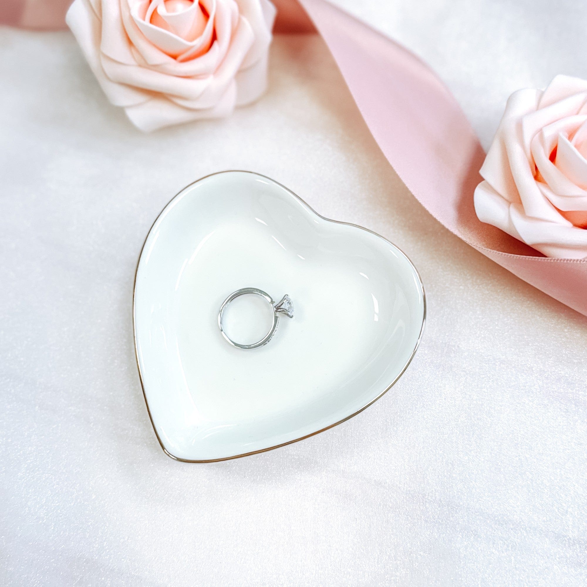 A white ceramic heart-shaped ring dish with a delicate gold rim, designed to safely hold rings or small trinkets for a wedding or bridal shower.