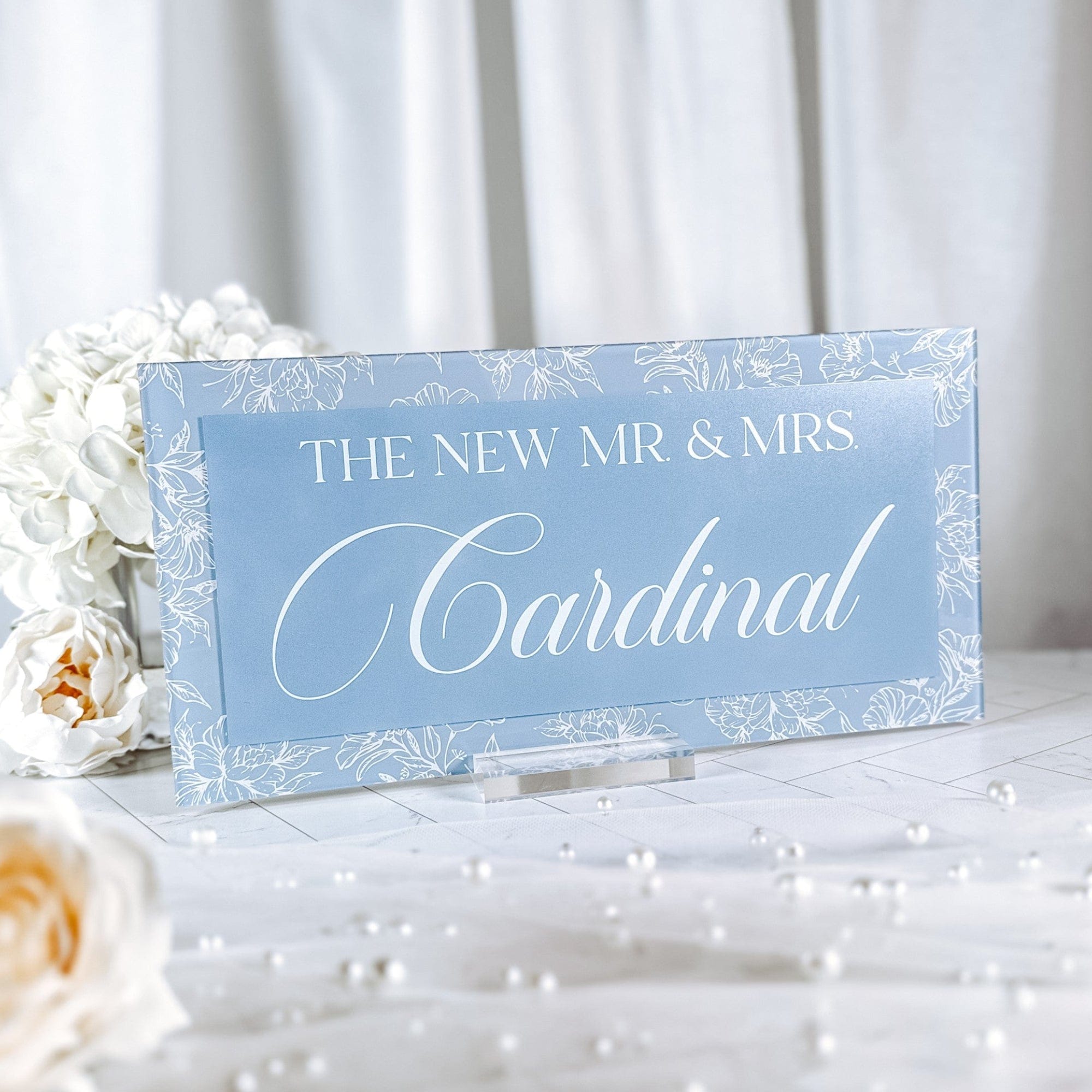 A personalized light blue acrylic wedding sign featuring white floral borders and a central text panel, displayed on a clear acrylic stand for reception decor.