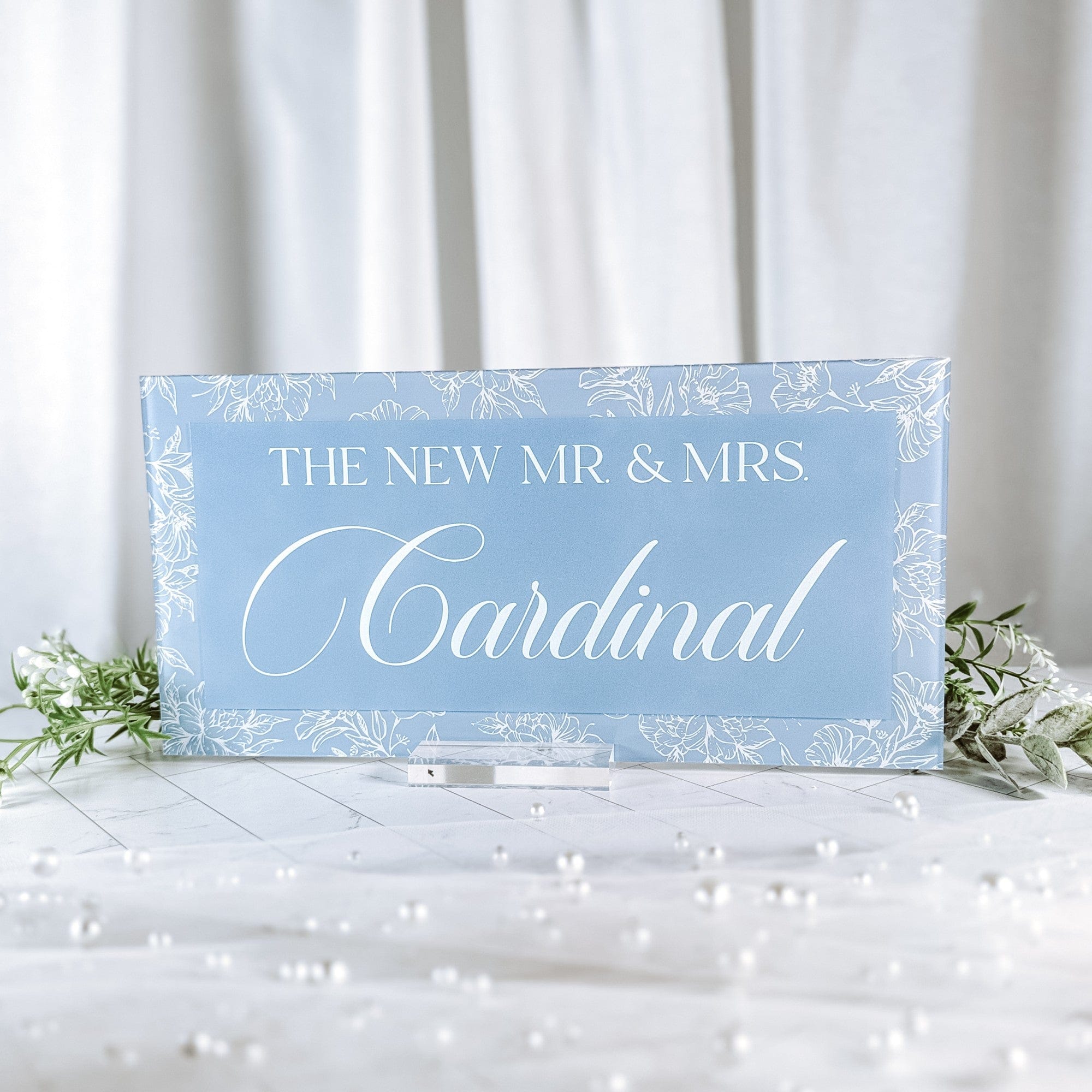 A rectangular light blue acrylic wedding reception sign features a delicate white floral line art border and is displayed on a clear stand, designed for personalized event decor.