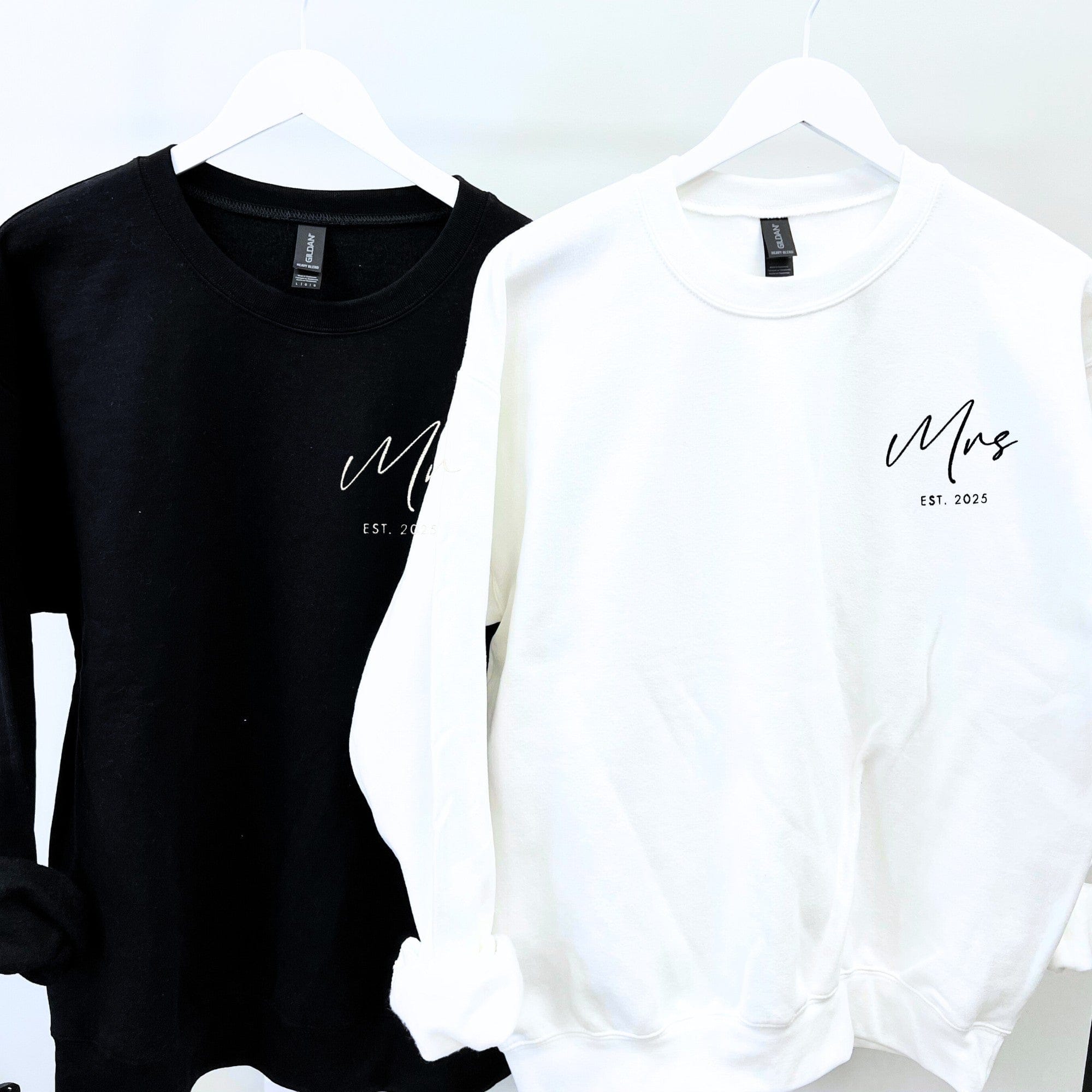 A pair of soft, personalized wedding sweatshirts, one black and one white, each featuring custom script lettering on the chest, hung on white hangers.