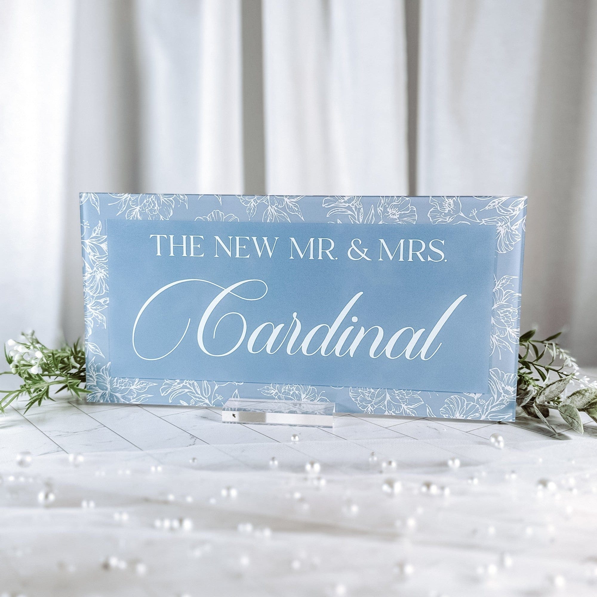 A rectangular light blue acrylic wedding table sign featuring an elegant white floral border design, presented on a clear stand with decorative greenery and scattered pearls.