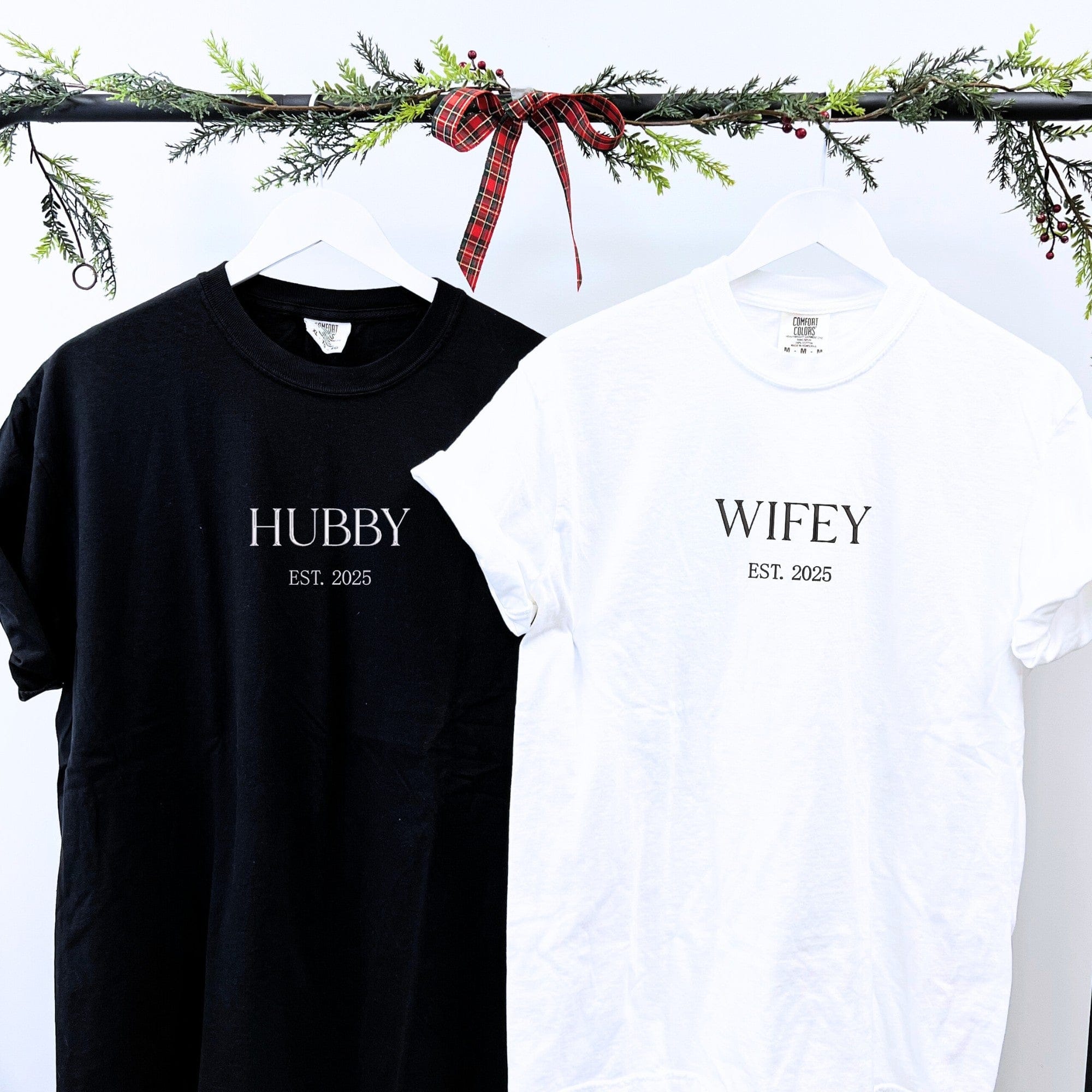 A black t-shirt and a white t-shirt, both soft cotton, designed as a matching set for a couple and featuring personalized text, hanging on a festive, garland-decorated rail.