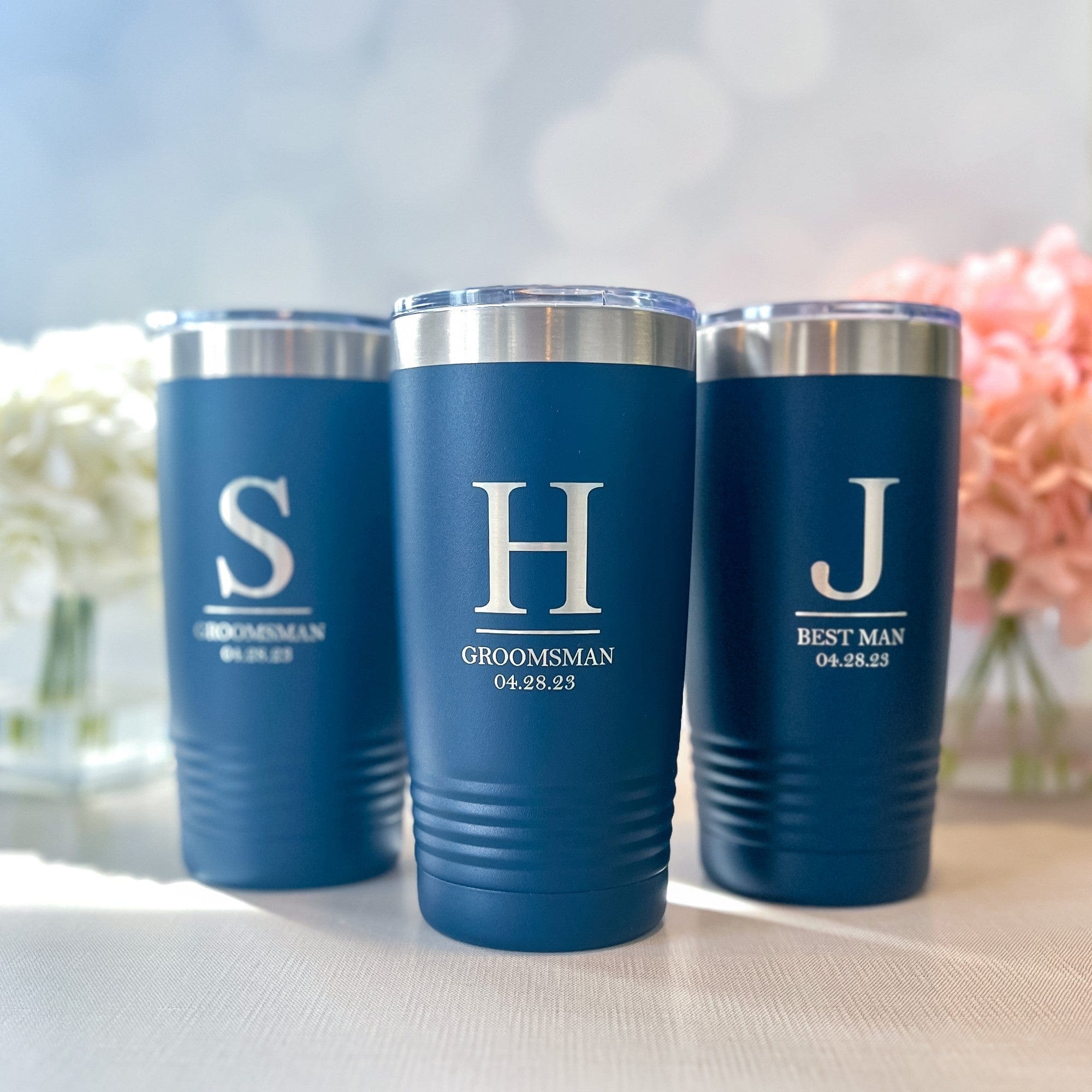 A set of three navy blue insulated metal tumblers with clear plastic and silver-toned lids, each customized with an initial and a wedding party role, serving as perfect wedding gifts.