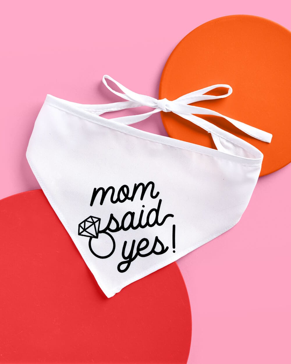 White pet bandana with "mom said yes!" text and diamond ring design, white ribbon ties, displayed on pink background