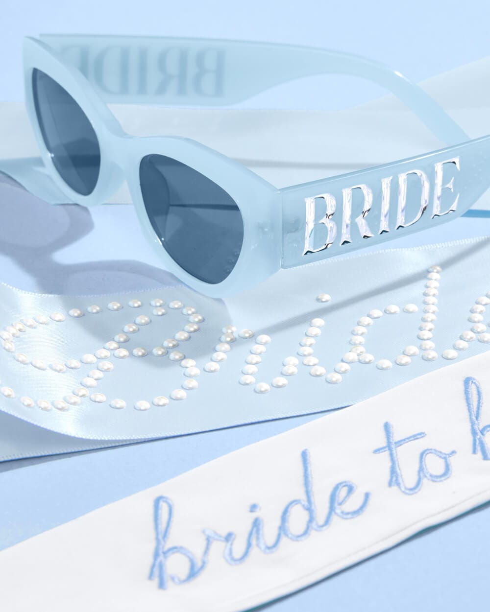 White oversized sunglasses with "BRIDE" text, pearl-studded blue sash, and blue "bride to be" hair bow on light blue background.