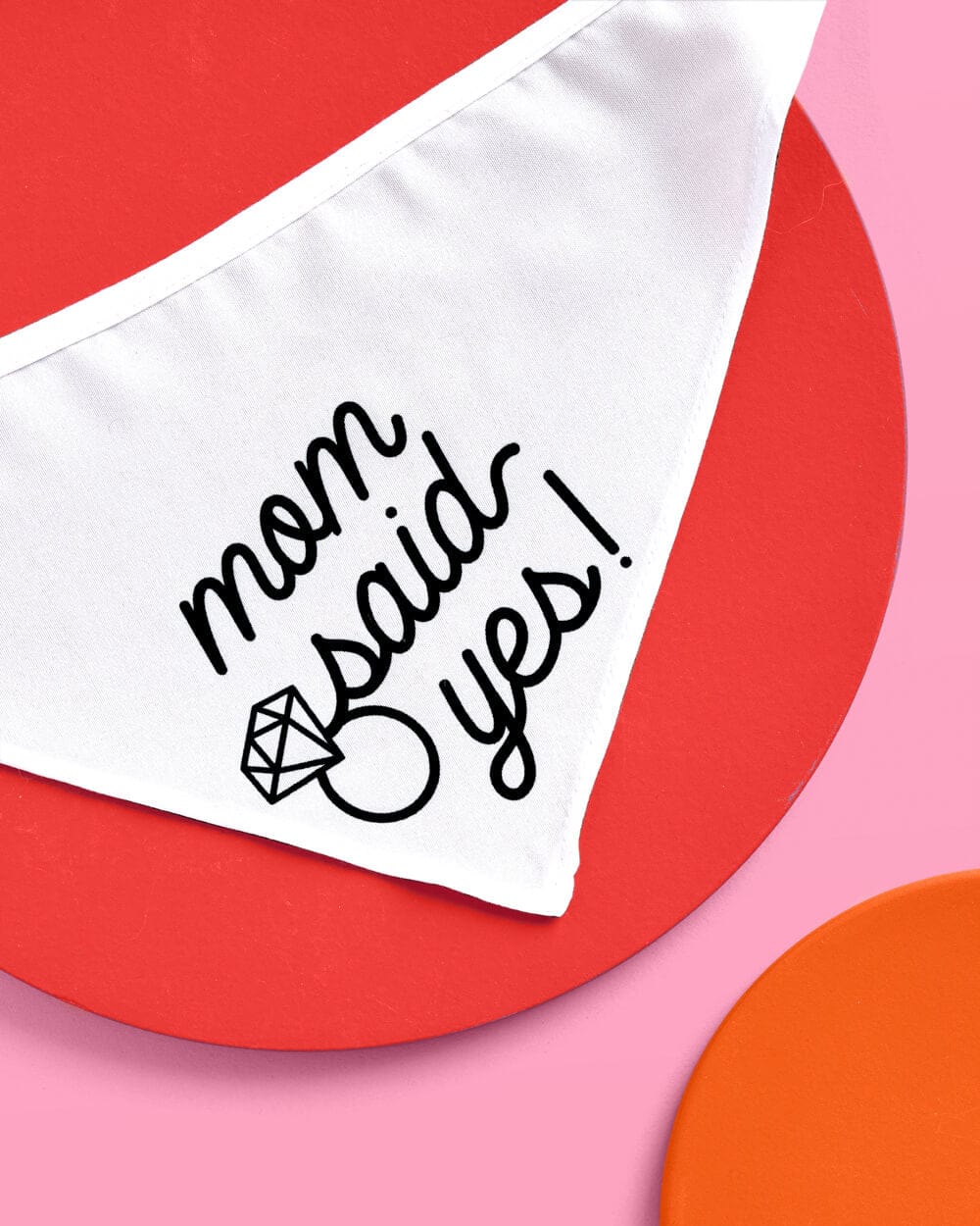 White pet bandana with "mom said yes!" text and diamond ring design in black handwriting on colorful background.