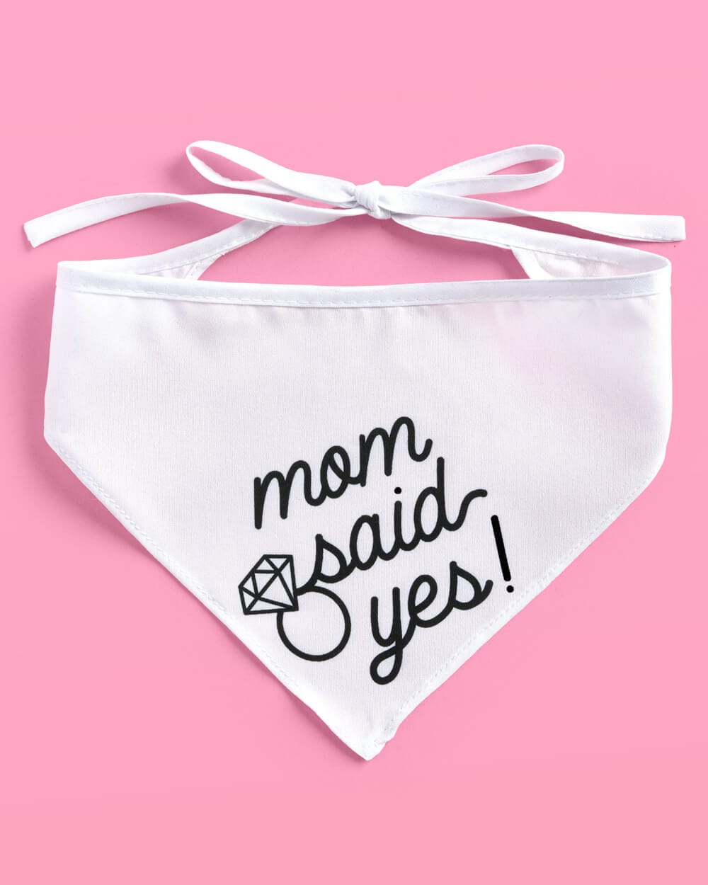 White pet bandana with "mom said yes!" text and diamond ring design, tied with white ribbons on pink background.