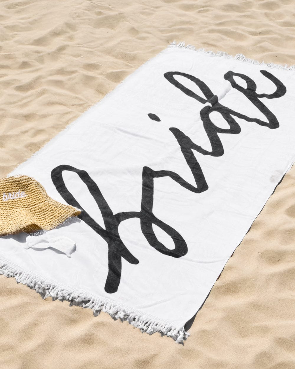 White cotton beach towel with "bride" text in black script, frayed edges, on sand