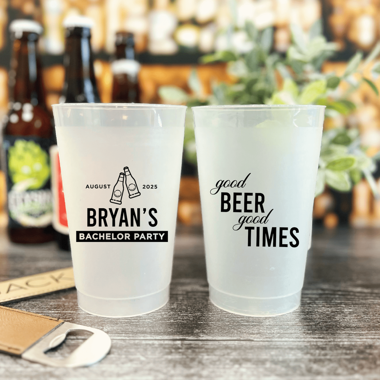 Frosted plastic cups with black text reading "Bryan's Bachelor Party August 2025" and "Good Beer Good Times" displayed on bar counter with beer bottles in background.