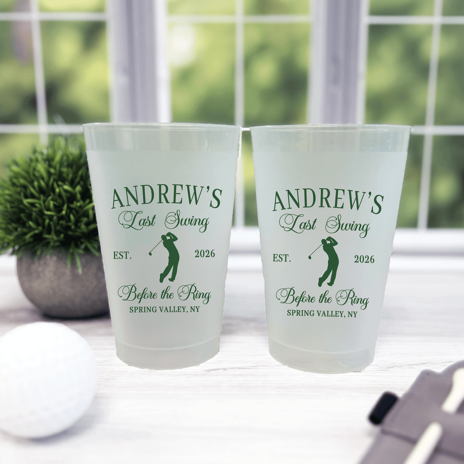 Frosted plastic cups personalized with "Andrew's Last Swing" and golf design, EST 2026, displayed on white table by window