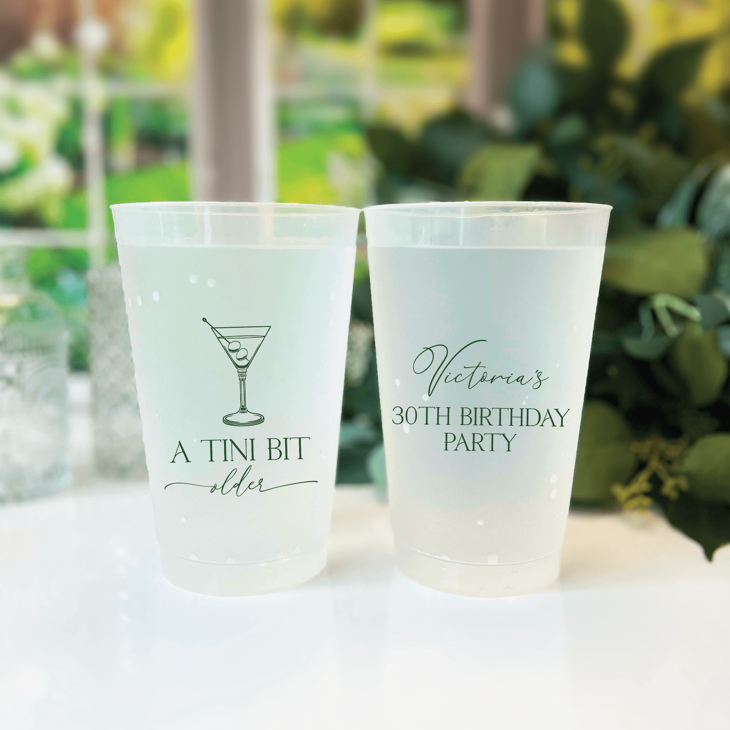 Two frosted plastic cups with custom text: left cup reads "A TINI BIT older" with martini glass illustration, right cup reads "Victoria's 30TH BIRTHDAY PARTY"