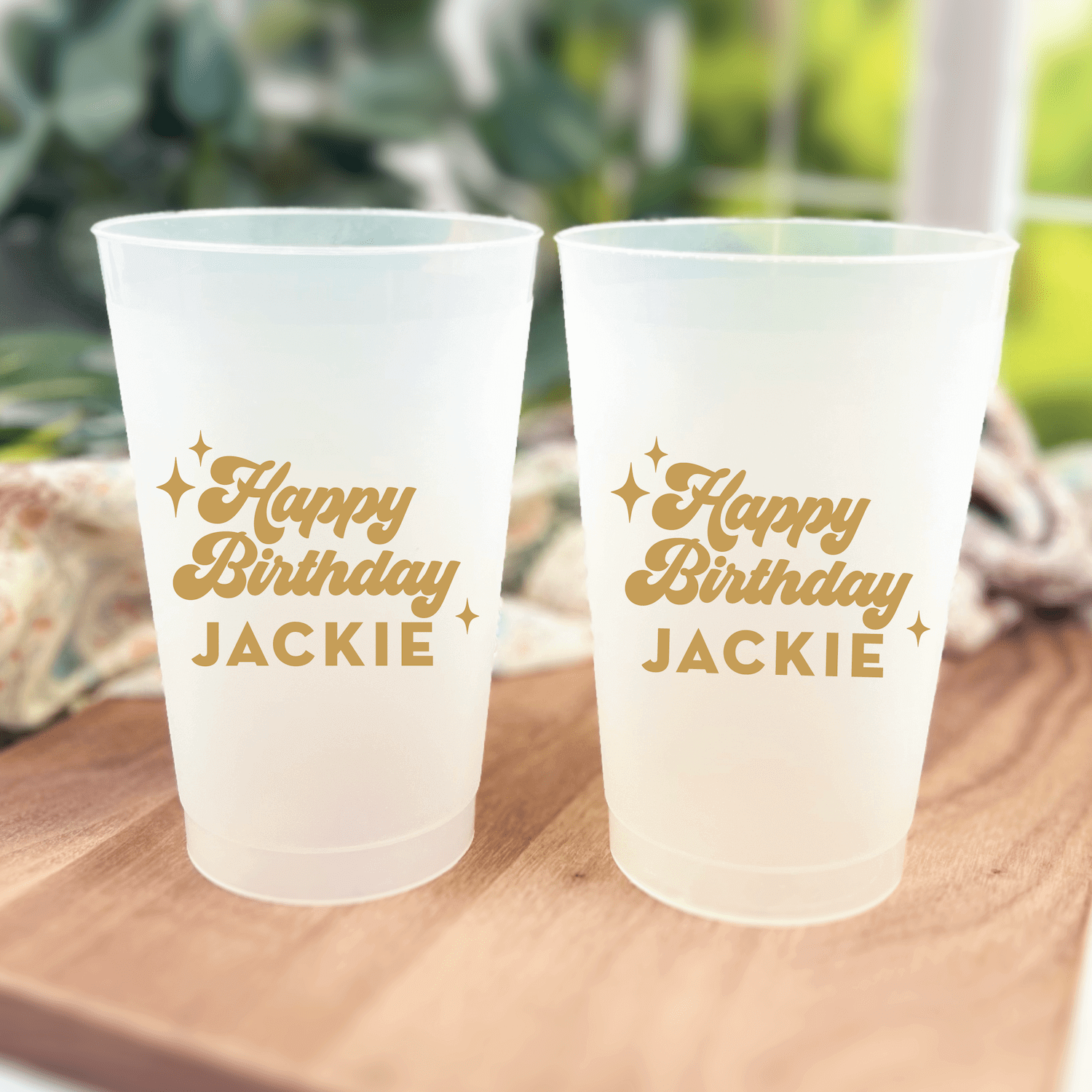 Two frosted plastic cups with gold "Happy Birthday JACKIE" text and star designs, displayed on a wooden surface outdoors.