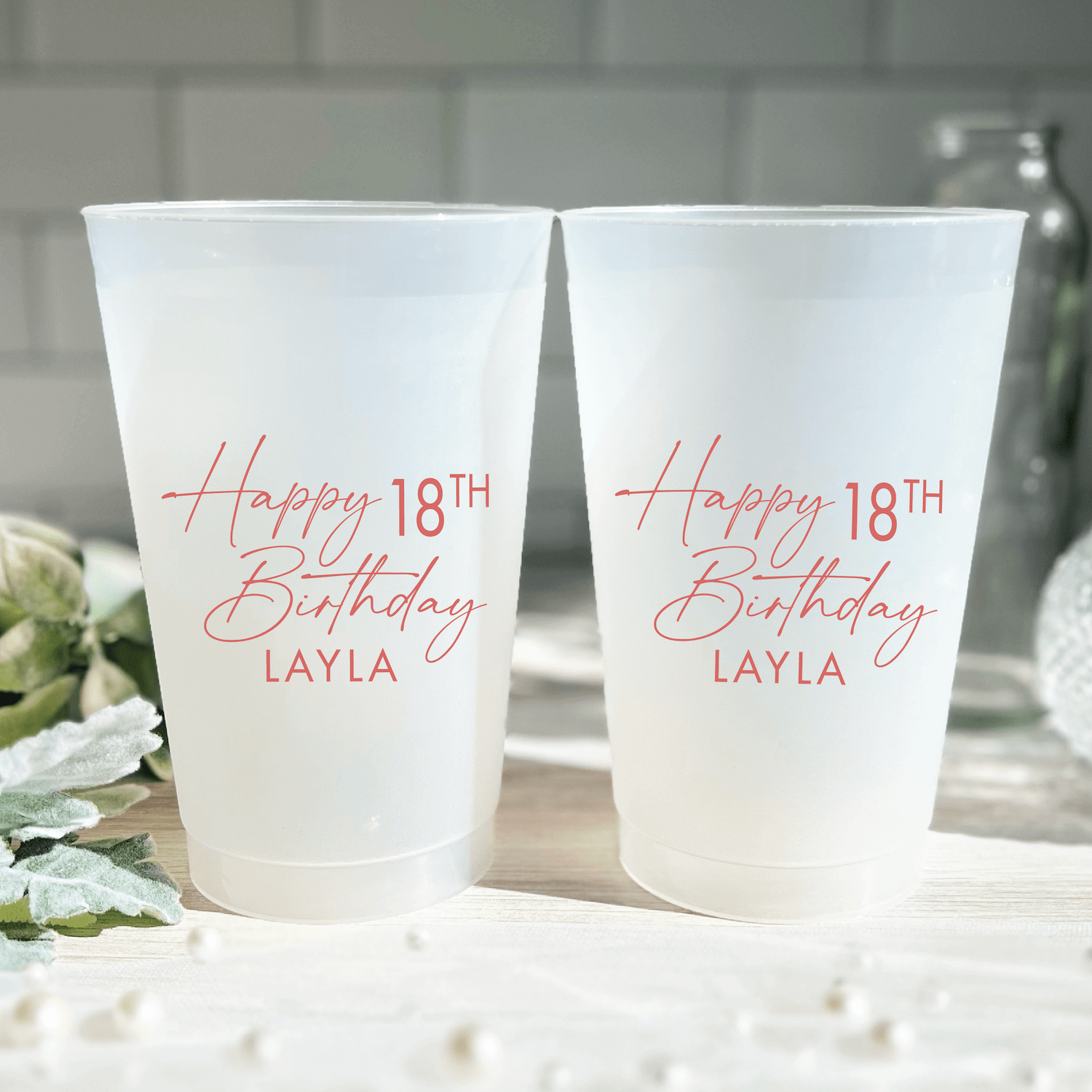 Two frosted plastic cups with pink text reading "Happy 18th Birthday Layla" displayed on white surface with flowers